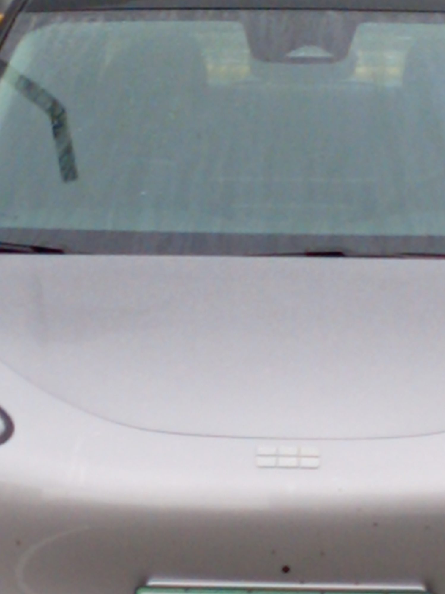 This image shows the front of a silver or light gray car with a windshield, windshield wiper, and what appears to be a sensor or camera housing near the top center of the windshield. The car has a rectangular emblem on the hood. The location and city are unknown from the provided image.