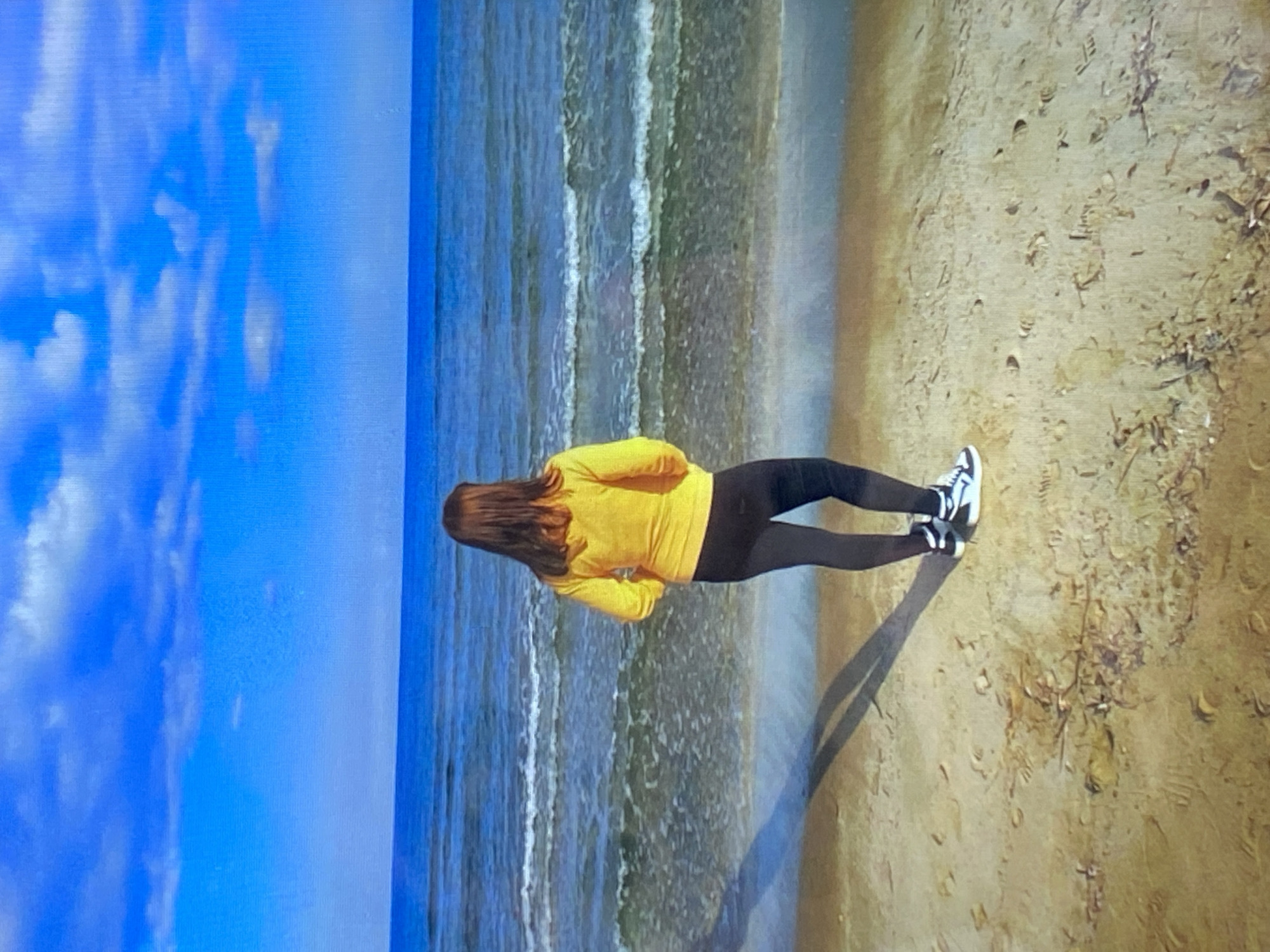 A person stands on a sandy beach facing a blue ocean under a bright blue sky with scattered white clouds. The person is wearing a yellow long-sleeved shirt and black leggings, with white and black sneakers. The waves are gently rolling onto the shore. There are footprints visible in the sand. The location is not specified in the image, nor is it possible to determine it is in the city of Pivdennoukrainsk, Ukraine.