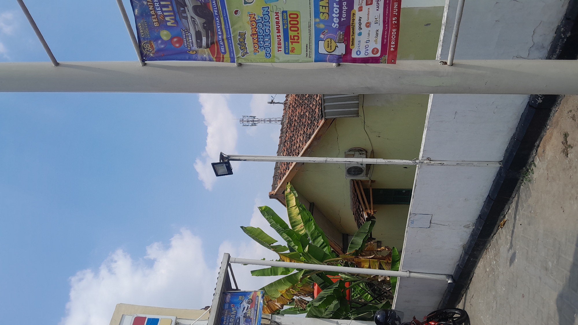 The image shows a street scene in Bangkle, Indonesia on a bright, sunny day with a clear blue sky and scattered white clouds. In the foreground, a white pole stands vertically, obscuring part of the view. To the left, a white wall with some visible cracks runs along the edge of a paved area. A red motorcycle is partially visible on the left edge of the frame.
Behind the wall, a light yellow building with a brown tiled roof is visible. An air conditioning unit is mounted on the side of this building. A cluster of large banana trees with green leaves is situated to the left of the building. A black garbage bin is located near the motorcycle.
On the right side of the image, several vertical banners are attached to poles. The top banner, featuring the number "38" and colorful balloons, advertises a promotion. Below it, a banner displays cartoon characters and text related to "POKÉMON SCHOOL COLLECTION" and "TEBUS MURAH Rp 15.000". Further down, another banner is partially visible, advertising "Sektor Tunai/Top Up" and various payment methods. A white pole runs vertically in front of these banners.
In the background, a cellular tower with multiple antennas is visible against the sky. The overall scene depicts an outdoor commercial or residential area.