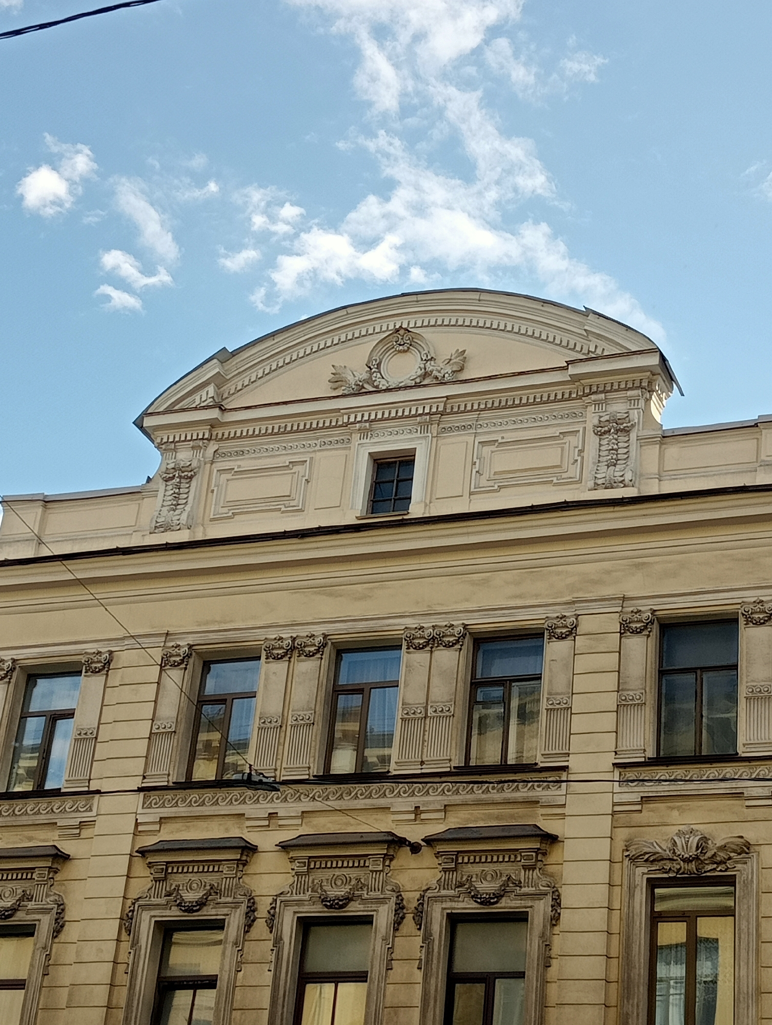 The image captures a section of the facade of a historic building in Saint Petersburg, Russia, under a bright blue sky with scattered white clouds. The building features a light yellow facade with classical architectural elements. The upper part of the building has a prominent arched pediment decorated with ornate stucco moldings, including a wreath and foliage. Below this, a band of intricate geometric patterns runs across the facade, punctuated by rectangular recessed panels. A small, dark rectangular window is centrally placed within the pediment area.
Further down, the building displays rows of windows. The upper row of windows is framed by decorative pilasters with ornate capitals and topped with projecting eaves. These windows are dark-paned and reflect the sky. A decorative frieze with a wave-like pattern runs below this row of windows. The lower row of windows is more elaborately decorated, featuring complex stucco surrounds with cartouches and floral motifs. These windows are also dark-paned and reflect the surrounding architecture and sky. Thin wires are visible strung across the facade, suggesting the presence of utility lines. The overall impression is one of stately, well-preserved classical architecture.