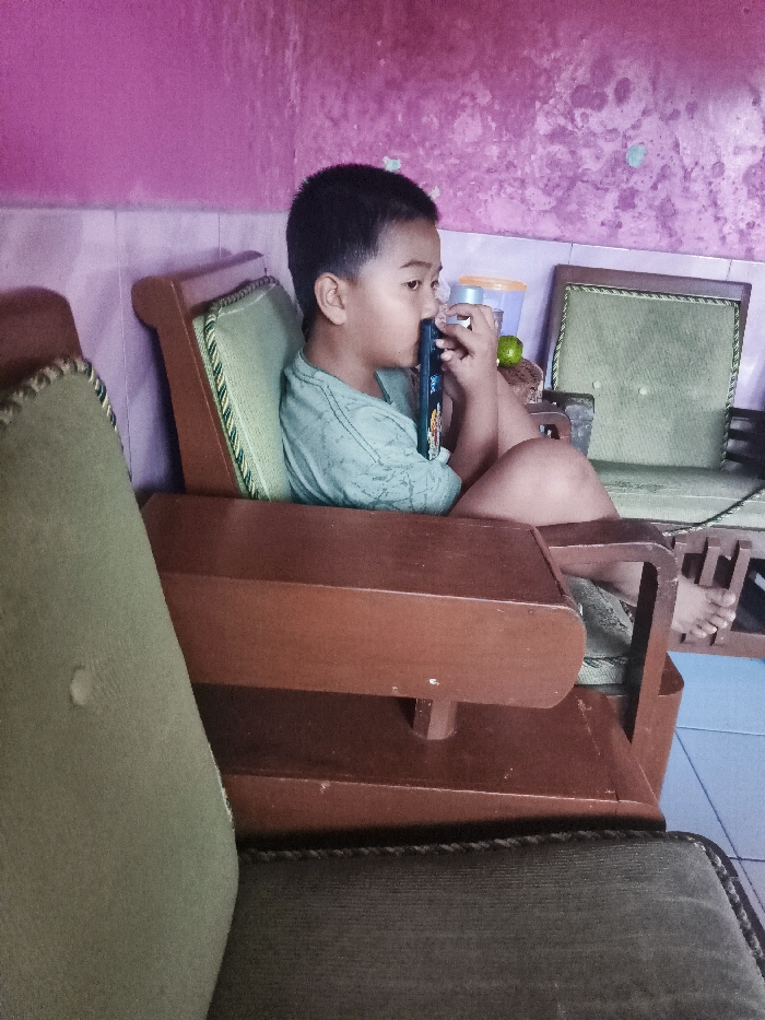The image shows a young boy sitting on a wooden armchair, facing the right side of the frame, in a home in Majalengka, Indonesia. The boy is holding a black phone to his ear and appears to be talking. His right foot is resting on the armrest of the chair. There is another wooden armchair behind him, to the right, with green fabric upholstery. The wall behind the chairs is pink with a textured finish.