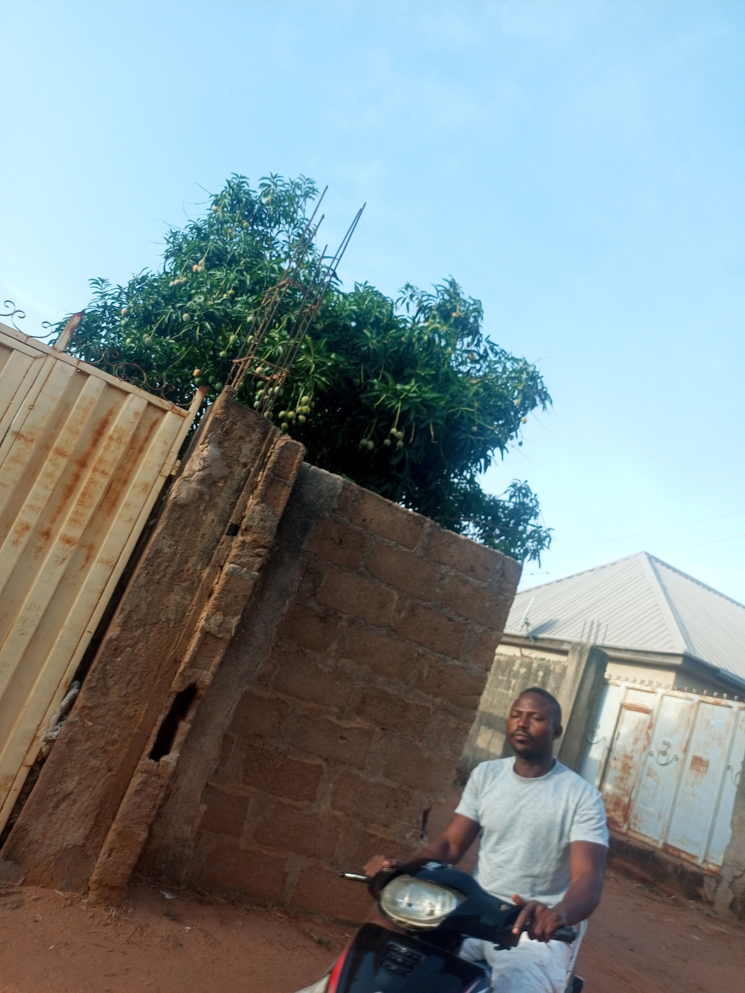 The image shows a man on a scooter near a wall in Tempure, Nigeria. The man is wearing a gray t-shirt and is facing forward. Behind him are a brick wall, a rusty metal gate, a tree with green fruit, and a building with a gray roof and a white metal gate. The ground is reddish-brown dirt. The sky is blue.