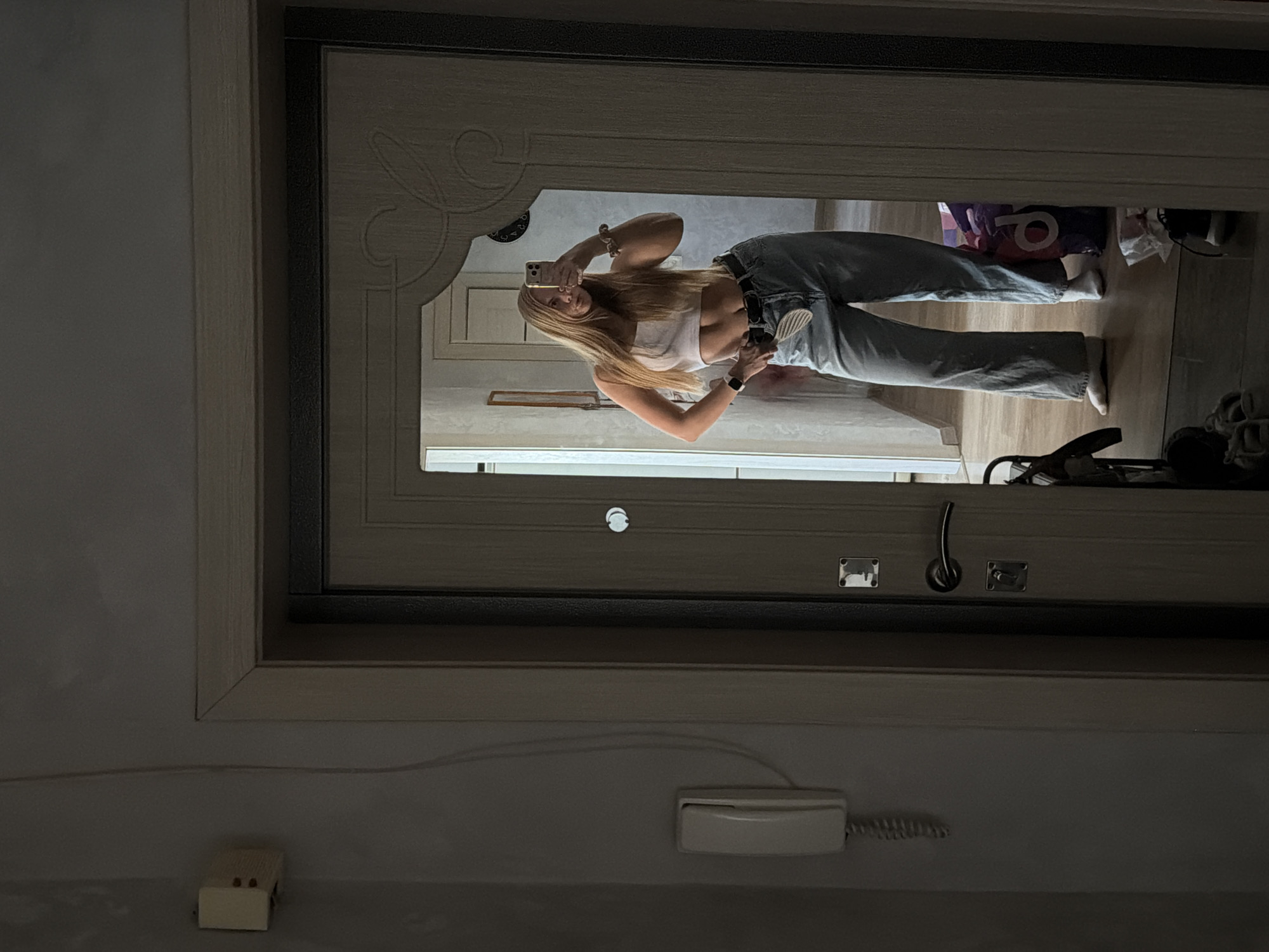 A reflection in a mirror shows a slender, fair-skinned woman with long blonde hair. She is wearing a white crop top, blue jeans, and a black belt. She is holding a smartphone in her right hand, presumably taking a photograph or video of herself. The mirror is mounted on a door with decorative carvings.
Behind the woman in the reflection, there are bags and other items on the floor. The room appears to be an entryway or hallway. Below the mirror, on a wall, is an intercom system. A small, rectangular object, possibly an electrical outlet, is visible near the bottom left corner of the image.
There is no verifiable information to support that this image was taken in Chaikovsky, Russia.