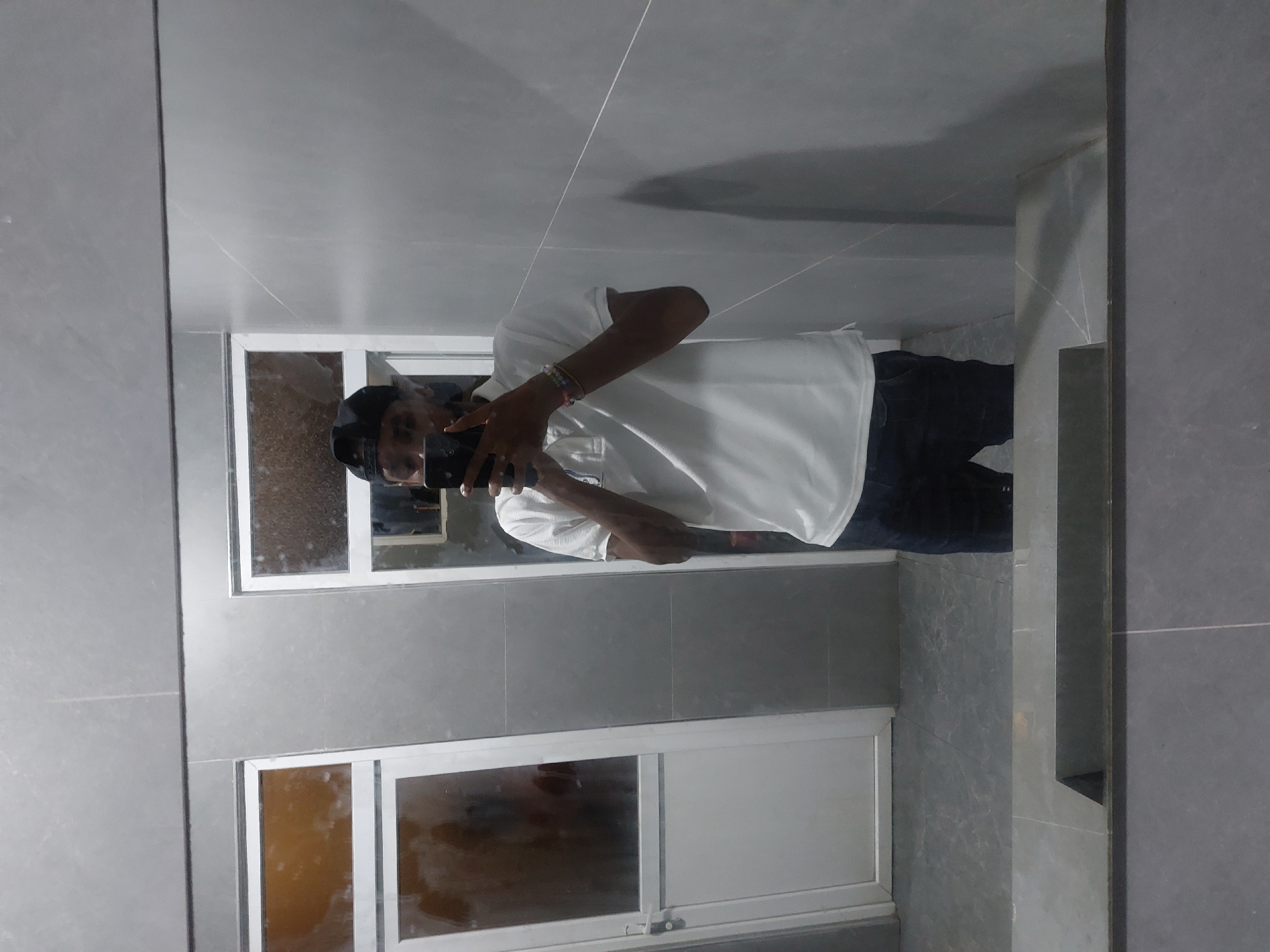 The scene depicts a male individual taking a mirror selfie in a modern, tiled interior space. The person, with dark skin, wears a black baseball cap reversed, a white short-sleeved polo shirt, and dark trousers. He holds a black smartphone horizontally with both hands in front of his face, capturing his reflection. Multiple colorful beaded bracelets are visible on his right wrist.
The environment consists of light grey, large-format tiled walls and a matching tiled floor. A dark grey, flat counter or integrated sink unit is visible in the foreground, at the base of the mirror's reflection. In the reflected background, two white-framed portals are present. The left portal appears to be a door with a handle and a translucent upper panel, reflecting a blurred, warm-toned interior. The right portal is a smaller, white-framed opening with frosted or textured glass.
This location is an indoor facility, likely a public or commercial restroom or changing area, consistent with modern architectural standards found in urban centers. The specific address or precise type of establishment (e.g., shopping mall, office building, hotel) within Dar es Salaam, Tanzania, is not discernible from the visual evidence.