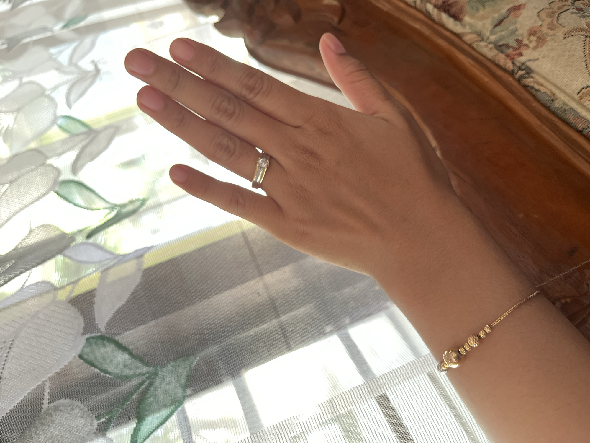 A close-up shot of a person's left hand and forearm is captured against a background of sheer curtains with a floral leaf pattern and a blurred wooden piece of furniture. The hand is positioned palm-up and angled towards the viewer, revealing a gold ring with a small, clear stone on the ring finger. A delicate gold bracelet adorns the forearm, featuring a series of small, rounded beads of varying sizes and textures. The skin tone is light to medium, and fingernails appear to be naturally finished or lightly polished. The lighting suggests an indoor setting with natural light filtering through the sheer curtains, creating a soft and slightly diffused illumination. The image does not contain any discernible elements that would indicate a specific location within Glagah Kulon, Indonesia.