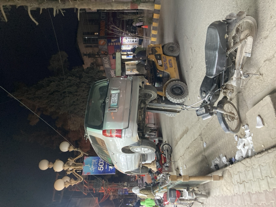 At night, a silver van bearing license plate BGT-764 is lifted by a yellow CAT Traffic Police Quetta forklift. A black motorcycle sits nearby. "Spring 50%" signage is visible next to the van. Background includes streetlights and building storefronts. "BRAND CITY" and "EPCOT" signs are readable. More motorcycles are parked on the street.
