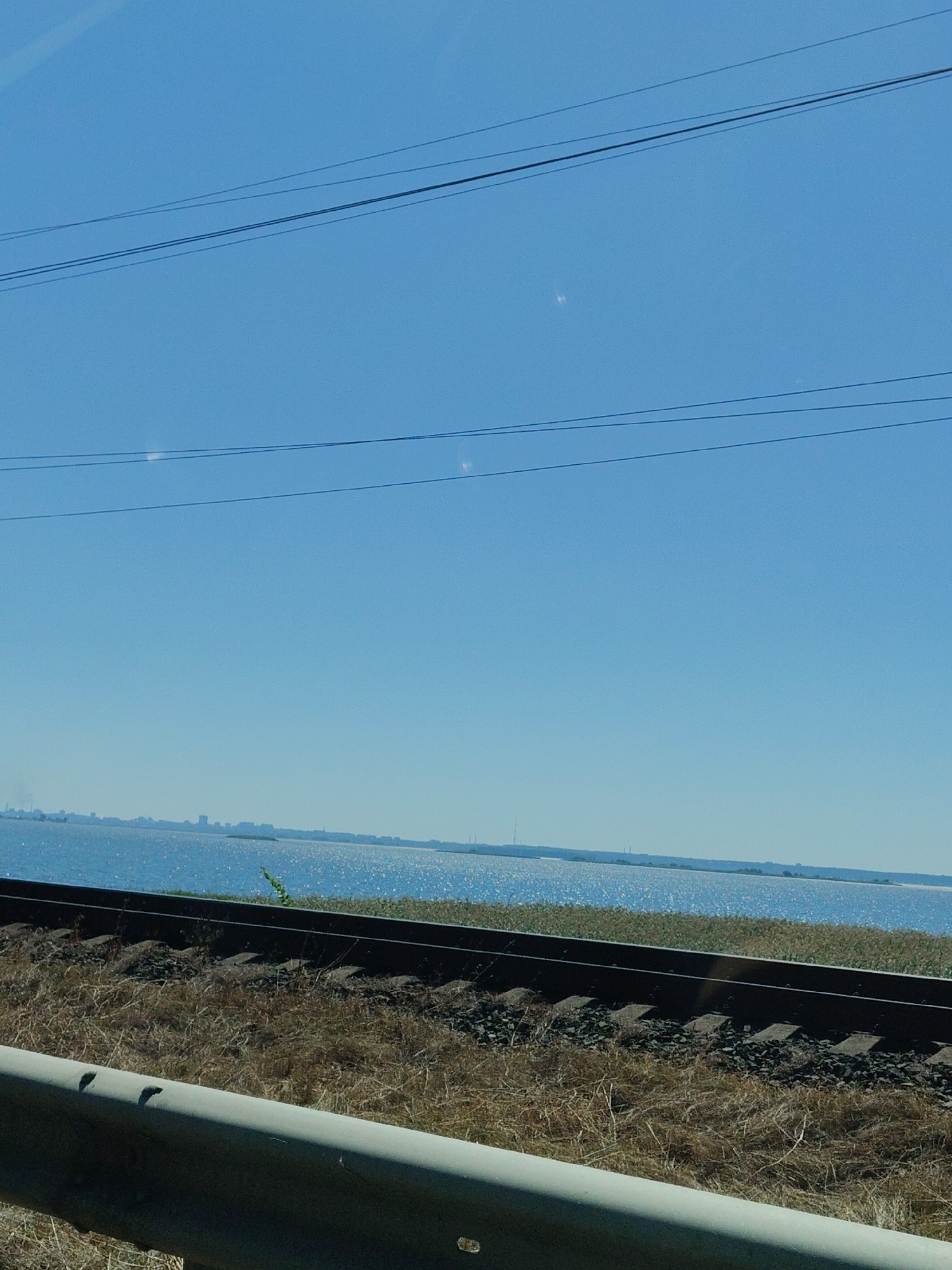 The image shows a view of a body of water with a distant city skyline, likely Blahodatne, Ukraine, under a clear blue sky. In the foreground, a railway track runs horizontally across the frame, bordered by dry grass and a concrete barrier. Several overhead power lines are visible against the sky. The water is shimmering with sunlight, and the distant city appears as a faint silhouette. There are no people or animals visible in the image.