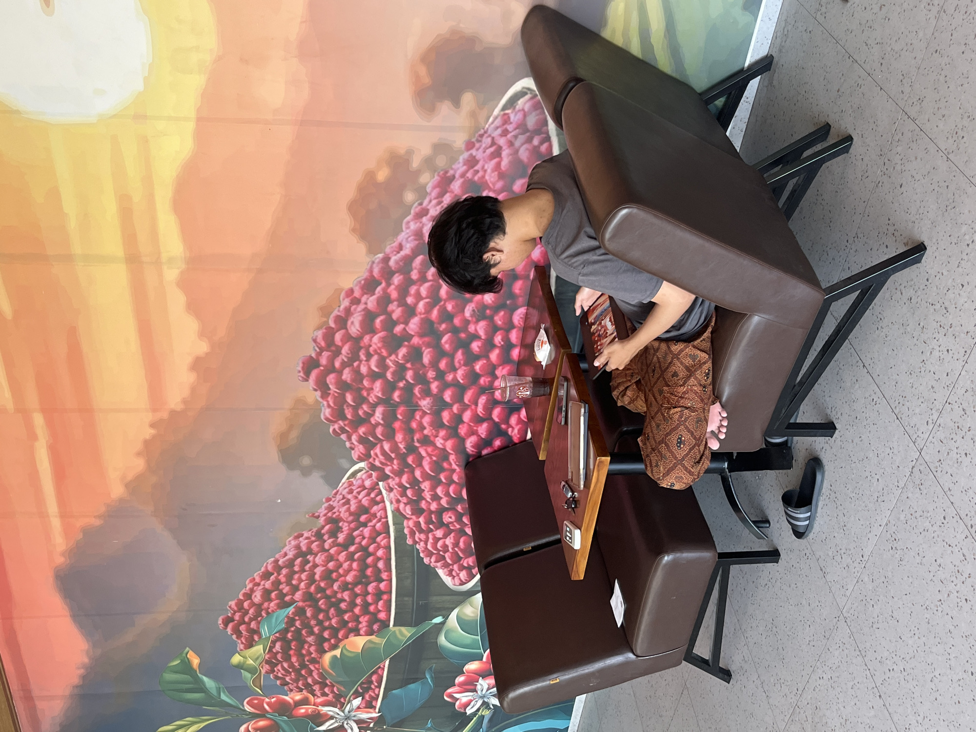 A young person sits in a brown leather booth in a cafe with a mural on the wall. The mural depicts a sunset over mountains and piles of red coffee beans. The person is looking at a tablet device and is wearing a grey t-shirt and patterned shorts. On the table are a drink, a tablet, keys, and what appear to be cards. A pair of black and white slides are on the floor. The cafe floor is tiled with grey speckles. The location is the Kuta Selatan area of Indonesia.