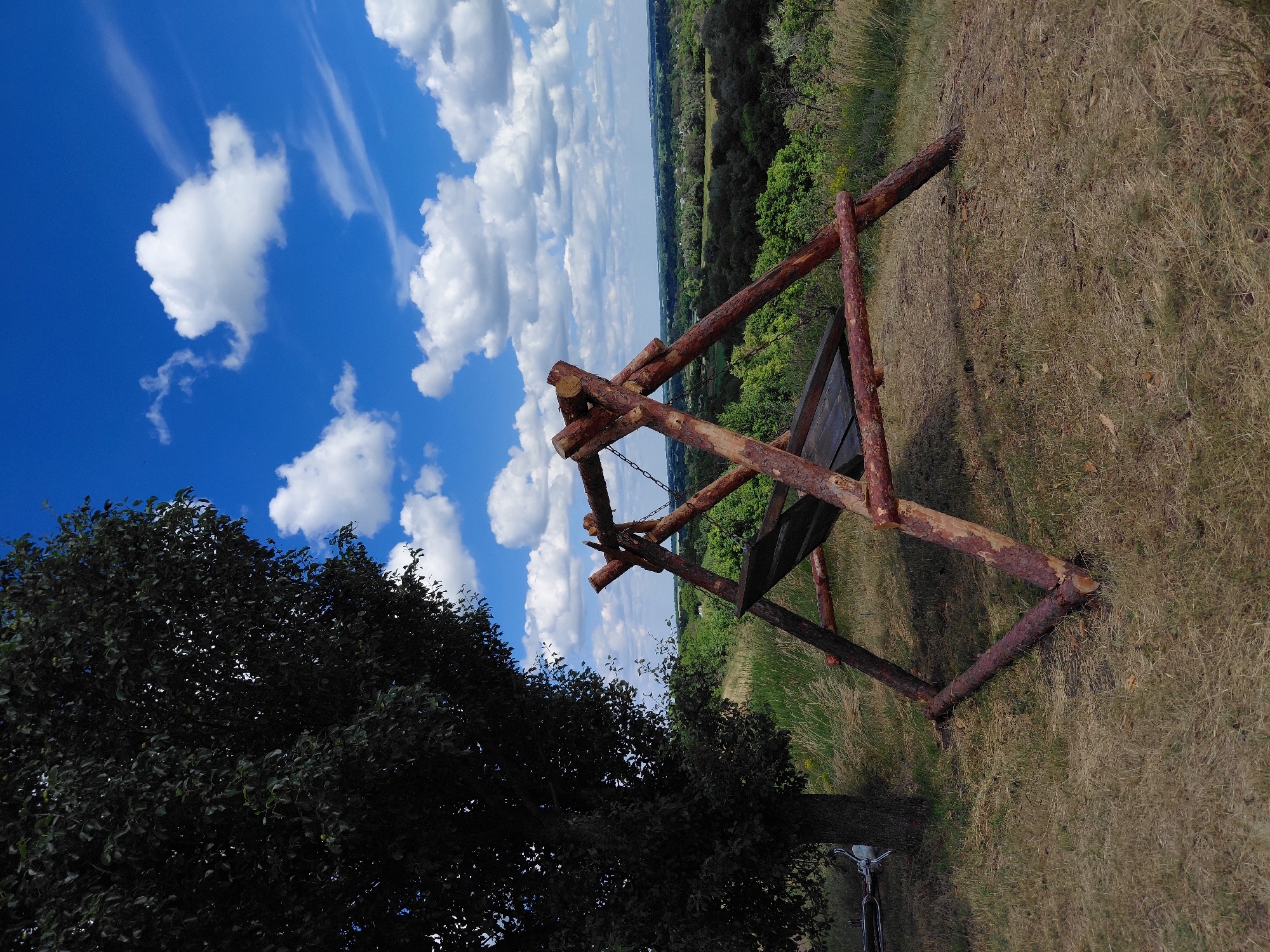 A wooden swing set is positioned on a grassy hilltop overlooking a valley on a sunny day in Ustyvytsia, Ukraine. The swing set is constructed from rough-hewn logs and has a wooden seat suspended by chains. To the left of the swing set, a large, dark-leaved tree stands. The ground is covered in dry grass and a few scattered leaves. The sky is bright blue with scattered white, puffy clouds. In the background, a lush green valley stretches out, dotted with trees and some buildings. A bicycle is visible lying on its side near the base of the tree. The scene appears peaceful and natural, typical of a rural landscape.