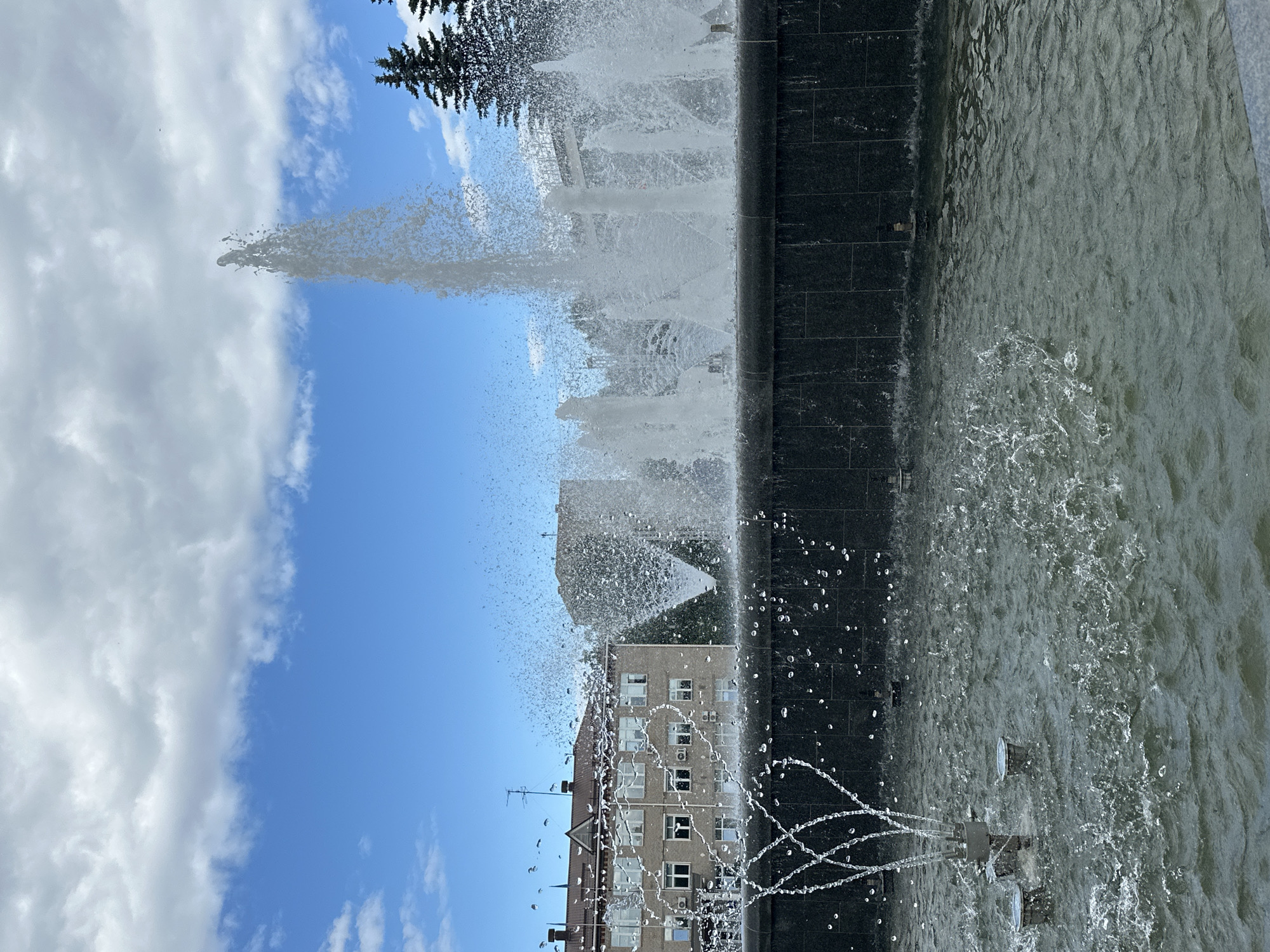 The image shows a public fountain in Chaikovsky, Russia, under a partially cloudy blue sky. The fountain has multiple jets of water spraying upwards and outwards, creating a dynamic and misty effect. The fountain's basin is lined with dark, polished stone. In the background, there are multi-story buildings with a light-colored facade and windows, typical of Soviet-era architecture. Evergreen trees are visible to the right of the fountain. The water in the fountain pool is rippled and shows splashes and droplets from the water jets. No people are visible in the image.