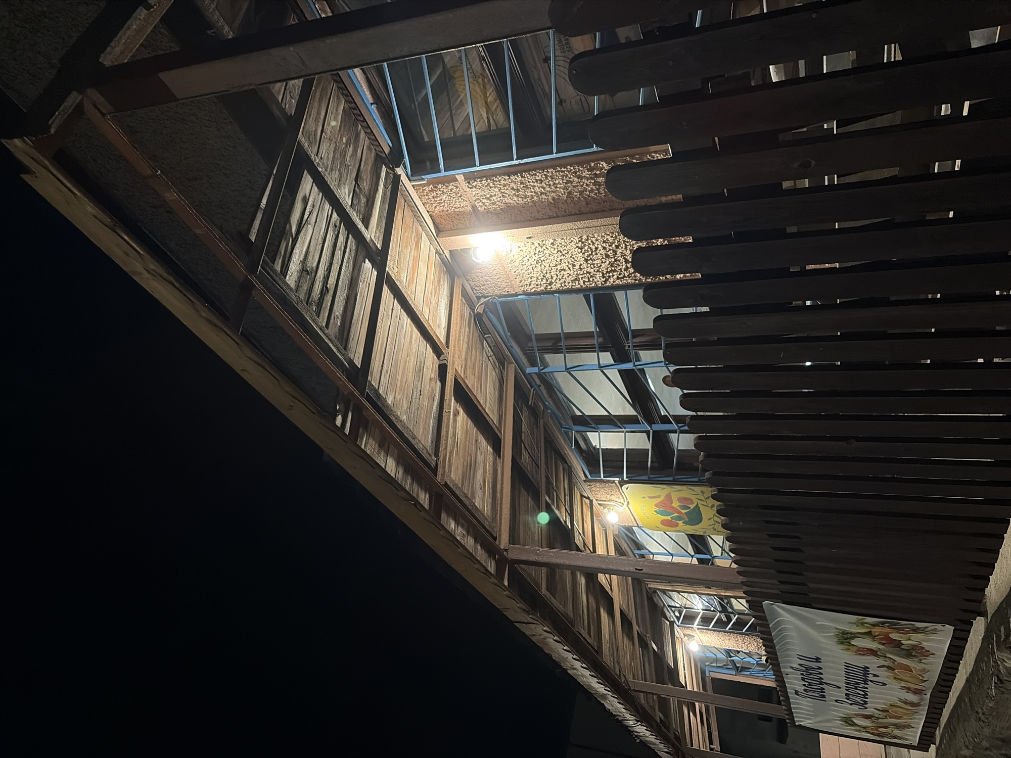 The image depicts a nighttime scene of a building's exterior, likely a market or shop, in the city of Obzor, Bulgaria. A wooden fence runs along the foreground. Behind the fence, a covered porch or walkway is visible, supported by metal and wooden beams. The roof of the porch is made of weathered wooden planks. Several windows with blue metal bars are present on the building's facade. Two light bulbs illuminate the porch area. A banner with images of fruits and vegetables and text in Cyrillic script, possibly indicating "Fruits and Vegetables," is displayed to the left, partially obscured by the fence. Another colorful banner with an image of produce is visible further down the porch. The overall impression is of a modest, open-air establishment at night.