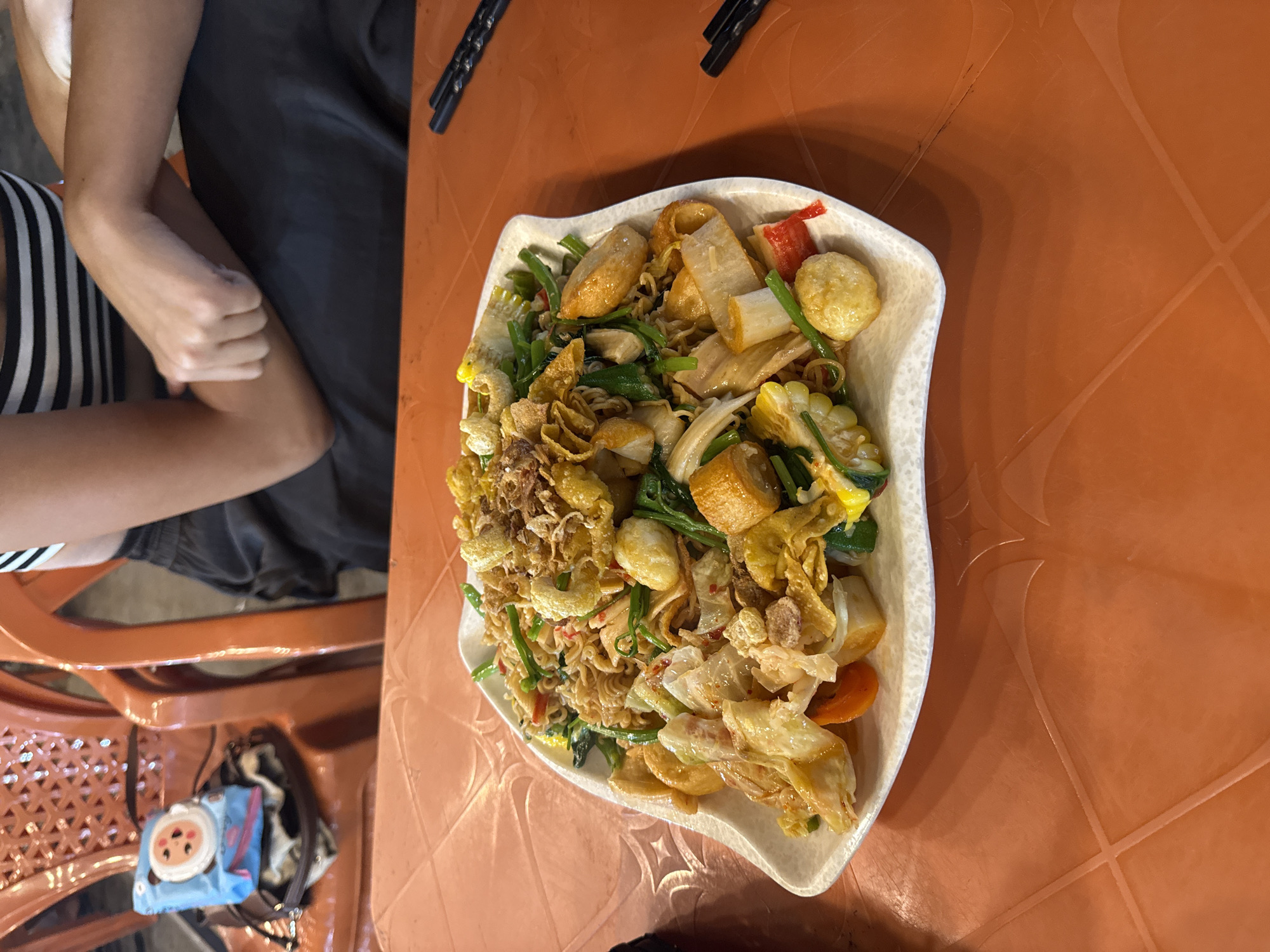 The image shows a plate of mixed food placed on an orange table with a geometric pattern. The food appears to be a Vietnamese-style mixed dish (possibly "mì trộn") containing noodles, vegetables (like water spinach), fish balls, fried onions, and other ingredients. Next to the plate are a pair of chopsticks. Seated next to the table are people. The scene likely takes place at a street food stall or a restaurant in Đà Nẵng, Vietnam, based on the food style and table setting.
