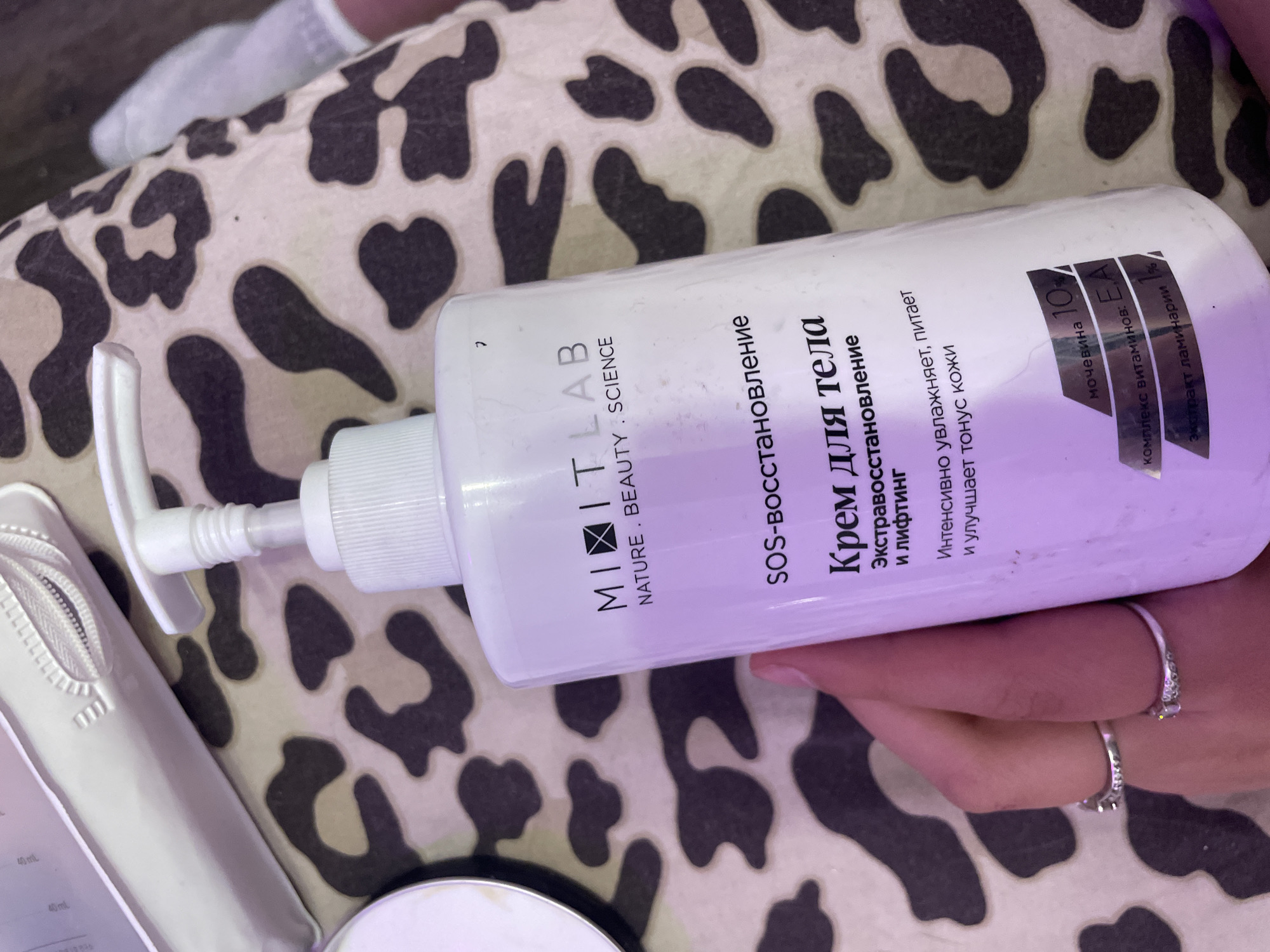 A hand with rings holds a white pump bottle of "MITIT LAB SOS-restoration body cream" on a leopard-print fabric. The bottle has Russian text indicating its purpose and ingredients. The background is a blurry close-up of the leopard print fabric and a portion of another white object, possibly a cosmetic bag. There is no visible indication of the city of Лесное Снт, Russia within the image.