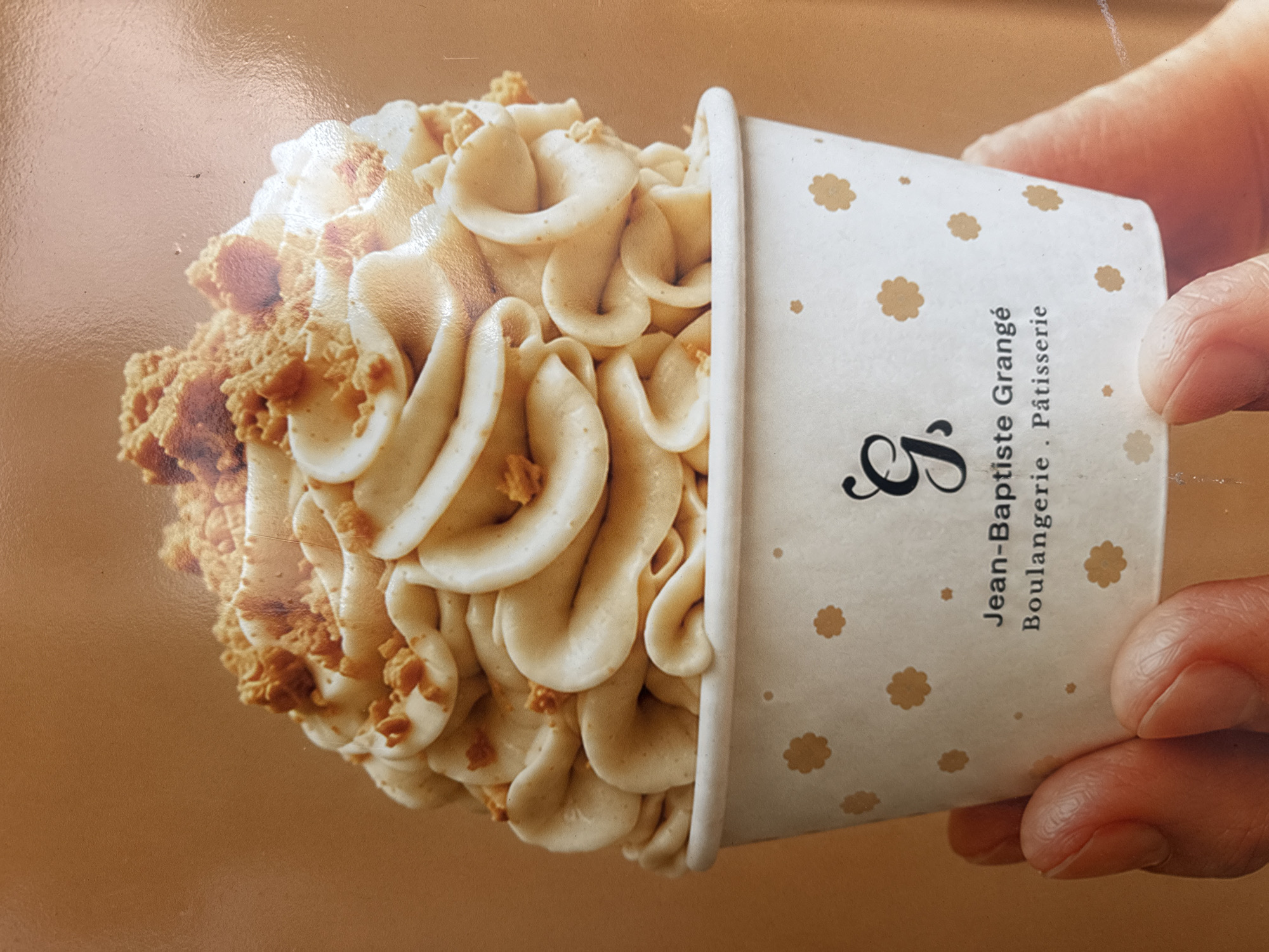 A close-up shot features a white paper cup filled with a swirl of light brown frosting, topped with crumbled cookie pieces. The cup is decorated with a pattern of small, light brown flower-like shapes and has text printed on it. The text reads "Jean-Baptiste Grangé Boulangerie Pâtisserie" with a stylized "G" above it. A person's hand is holding the cup from the side, with fingers visible around the bottom. The background is a plain, warm brown color. The scene is consistent with the display of a dessert item from a bakery.