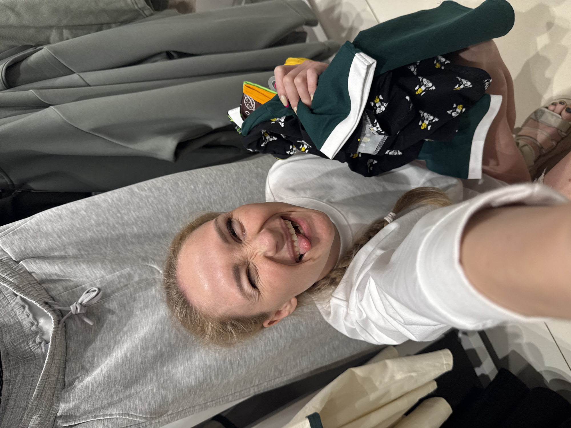 A woman is taking a selfie indoors. She is holding a stack of clothing items, including green shorts with a white stripe, and black shorts with a white cow pattern. She is wearing a white shirt and light-colored pants. The background includes a rack of gray clothing. There is no indication of the location being Chaikovsky, Russia.
