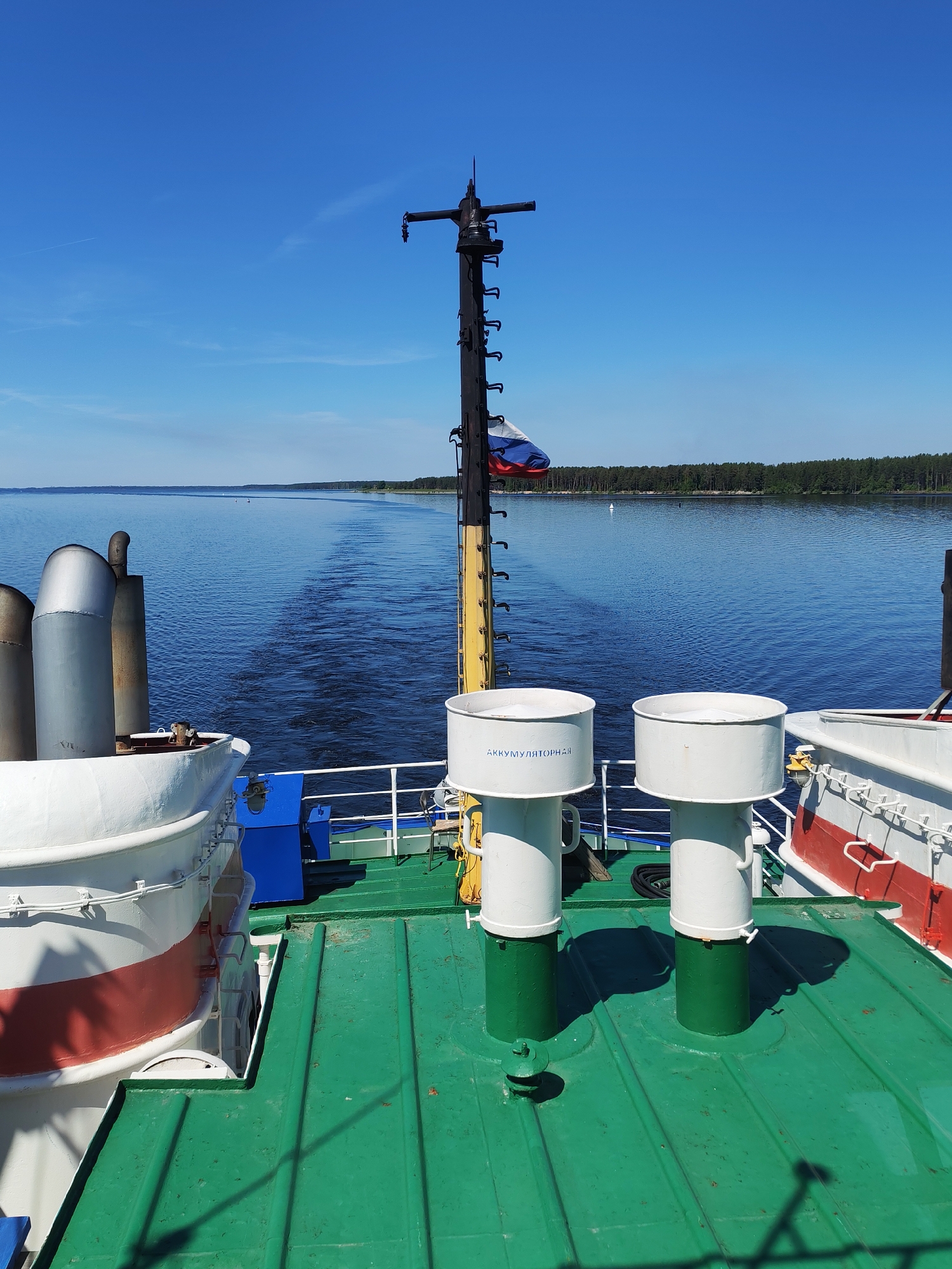 This image depicts the stern view from a vessel traversing a wide body of water, identified as being in the vicinity of Kostino, Russia. The sky is clear and brilliant blue with minimal cloud cover, exhibiting a faint white contrail in the upper left quadrant. The water surface is calm and deep blue, displaying a distinct wake directly behind the vessel, indicating forward motion.
In the immediate foreground, the vessel's deck is visible, primarily green in color with some visible wear. On the left side, three grey cylindrical exhaust pipes or vents are partially seen. Adjacent to these, a white lifeboat with a red stripe is visible. Centrally positioned on the deck are two prominent white cylindrical ventilator cowls with green bases. The left cowl bears the blue Cyrillic inscription "АККУМУЛЯТОРНАЯ" (AKKUMULYATORNAYA), signifying 'Battery Room' or 'Accumulator'. On the right, another white structure with a red stripe, likely part of the vessel's superstructure or another life-saving device, is partially present. Blue and white railings run along the perimeter of the deck.
A tall, dark-colored mast with a yellow base stands centrally in the midground, extending upwards and fitted with various components resembling steps or a ladder. A Russian flag, characterized by horizontal white, blue, and red stripes, is affixed to the mast and is visible fluttering. In the distance across the water, a solid line of dense green forest forms the far bank, extending across the horizon. A white buoy is faintly discernible on the water closer to the forested bank. No individuals are visible in the scene.