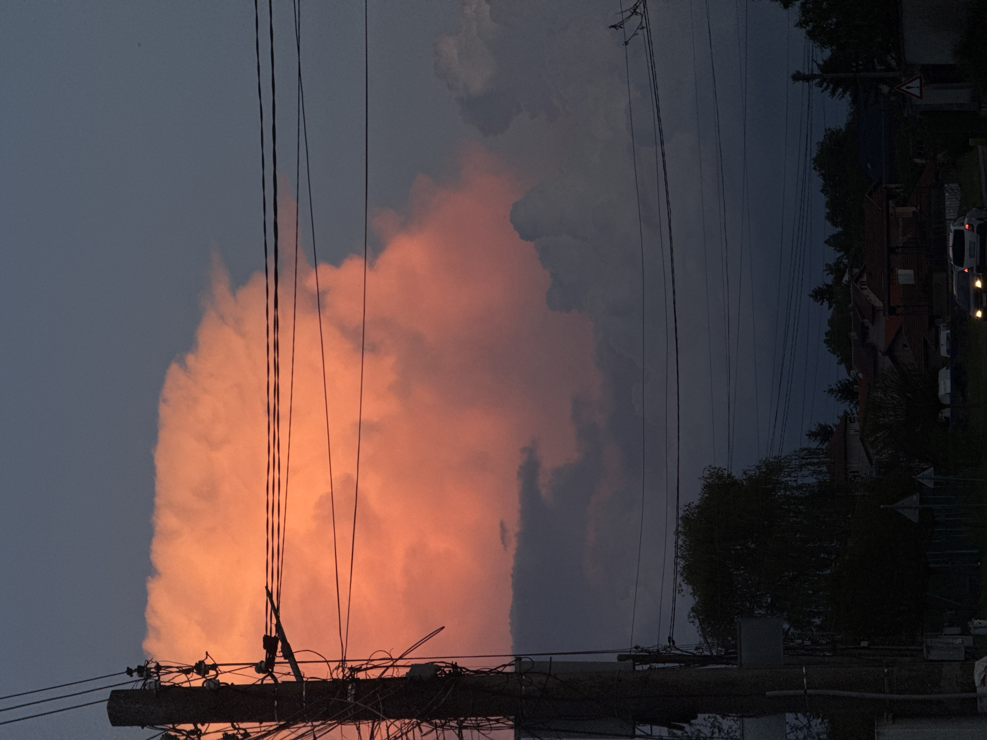 The image depicts a late evening scene in Petarch, Bulgaria. In the foreground, an electrical pole with numerous wires running overhead is visible. To the left of the pole, a large, orange-lit cloud dominates the sky, contrasting sharply with the darkening blue of the sky. To the right, the scene transitions into a residential area, with houses and trees silhouetted against the dusk. A few vehicles with headlights on are visible, suggesting street activity. The overall scene indicates a moment of transition between day and night in a suburban or residential environment.