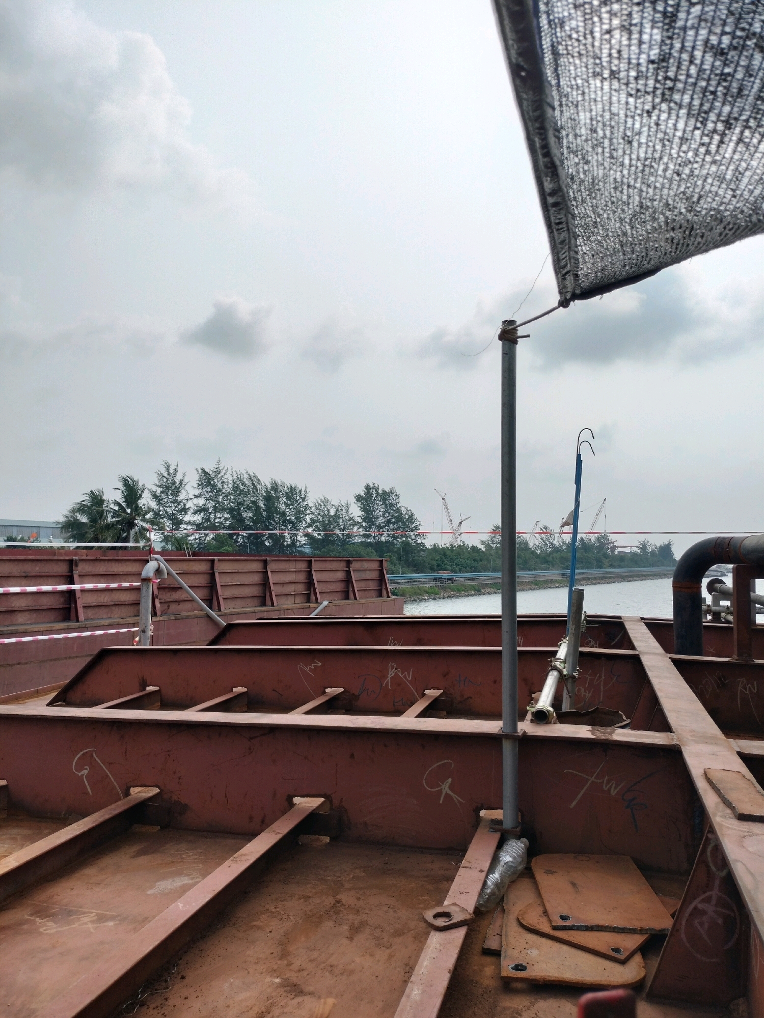 The image shows the interior of a large, partially constructed metal structure, possibly a ship or industrial platform, in Batam, Indonesia. The structure is made of thick, rusty red metal beams and plates, arranged in a grid-like pattern. Some of the metal surfaces have graffiti or markings. In the foreground, there are various metal components, including some flat plates stacked on top of each other. A metal pole with a piece of netting attached to its top stands vertically in the center of the frame. A plastic bottle is visible at the base of the pole.
In the background, a body of water is visible, with a line of trees on the opposite bank. Beyond the trees, there are several construction cranes, suggesting an industrial or port area. A road and some buildings can also be seen in the distance. The sky is overcast with clouds. The overall impression is of an industrial construction site, likely in a port or shipyard setting in Batam, Indonesia.