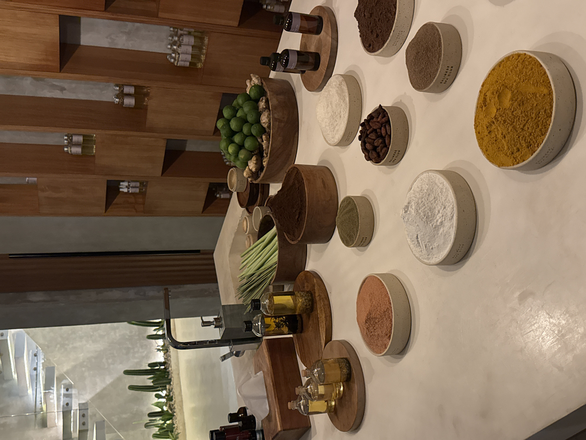 The image shows a display of natural spa ingredients, likely in a shop or spa, possibly located in Tibubeneng, Indonesia. On a white surface are arranged bowls and wooden platforms holding various substances, including brown cocoa powder, yellow turmeric powder, a brown spice mix, lemongrass, lemons, essential oils, and brown chocolate beans. In the background, a wooden shelving unit displays rows of small, clear bottles. A modern sink area with a dispenser and green plants is also visible.