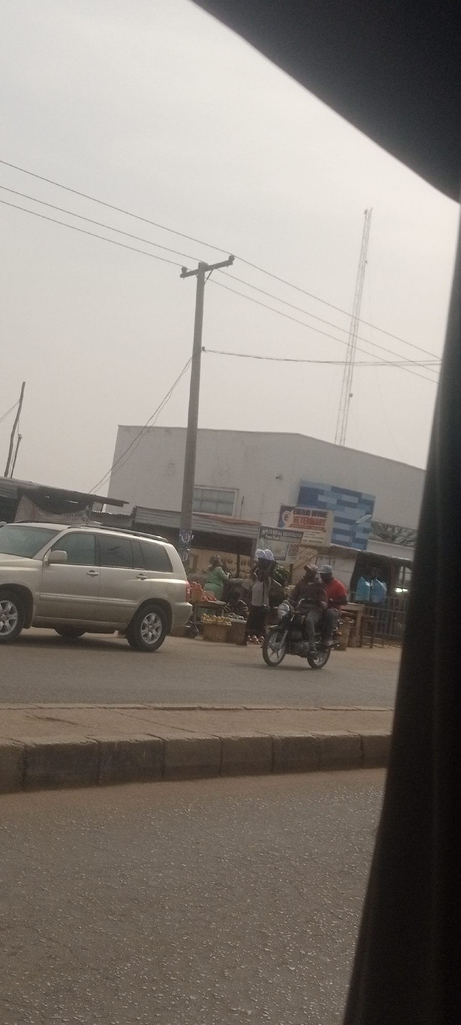 This image captures a street scene in Sabon Tasha, Nigeria, under a hazy, overcast sky, indicating daytime. The view is partially obscured on the right side by a dark object, likely part of a vehicle's interior.
In the foreground, an asphalt road with a concrete curb is visible. A light-colored SUV, possibly a Toyota Highlander, is positioned on the left side of the road, facing right. To its right, a motorcycle with two male riders is moving along the road; one rider wears a red shirt, and the other, in a darker shirt, wears a light-colored cap or helmet.
Behind the vehicles, a vibrant street life unfolds with several people gathered around what appear to be market stalls laden with produce, including fruits and vegetables. These activities take place in front of a series of buildings. A tall utility pole with numerous power lines stands centrally, connecting to other poles and disappearing into the hazy sky.
In the background, a prominent white building features a distinctive blue and white patterned section on its right side. A large sign on this building clearly displays the text "EKEKU DIVINE VETERINARY." Below it, partially obscured, is "133 ROYAL BENDOBONG" (the last word's spelling is slightly uncertain due to blur). Above the buildings, a tall, slender communication mast or antenna is visible against the muted sky. The overall scene depicts a bustling, functional urban or semi-urban environment.
