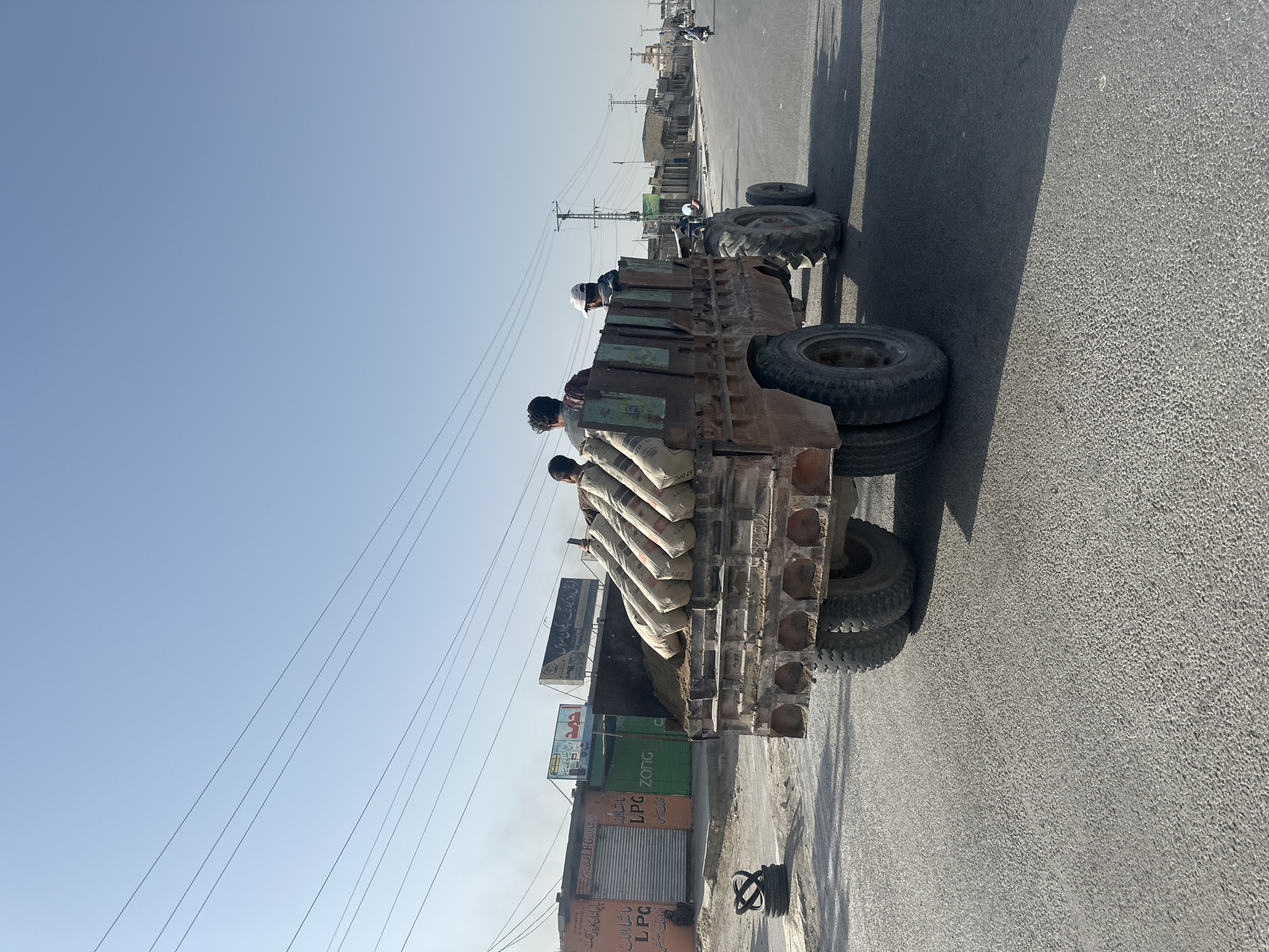 The image shows a flatbed truck moving along a street in Quetta City Tehsil, Pakistan. The truck is loaded with several bags of cement, and three individuals are riding atop the cargo. The truck appears old and well-used. The street is paved, and buildings line the sides of the road, including one with signage in Urdu. Power lines are visible overhead against a clear sky. Another vehicle is visible further down the road.
