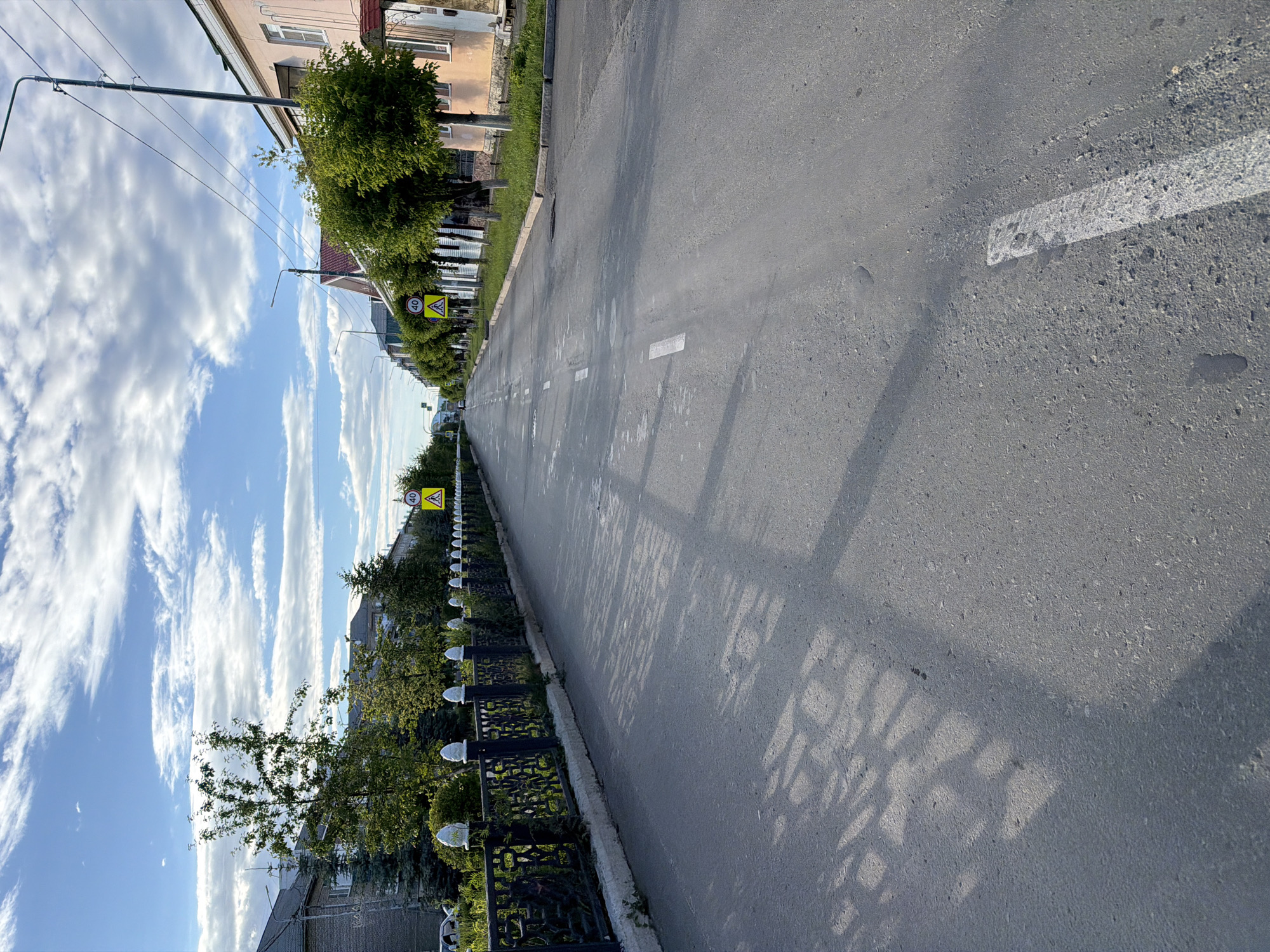 The image depicts an asphalt road with white markings, stretching into the distance. On the left side of the road, there's a sidewalk bordered by a decorative metal fence with white lights. Beyond the fence, trees and houses are visible. The sky is a bright blue with dramatic white clouds, suggesting a sunny day with some wind. A road sign indicating a speed limit of 40 is visible. The scene appears to be a residential street, possibly in a town or village. The perspective is from ground level, looking down the road. The light suggests it is daytime, likely late afternoon due to the long shadows cast on the road by the fence. There are no people visible in the image. The overall mood is serene and ordinary, capturing a quiet street scene.