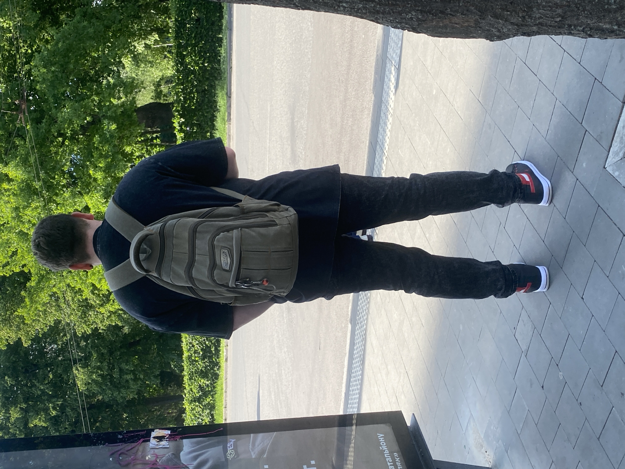 A man stands on a sidewalk in Poltava, Ukraine, wearing a black t-shirt, black jeans, and red and black shoes. He has a khaki-colored backpack. He is looking at an advertising kiosk. Trees are visible in the background.