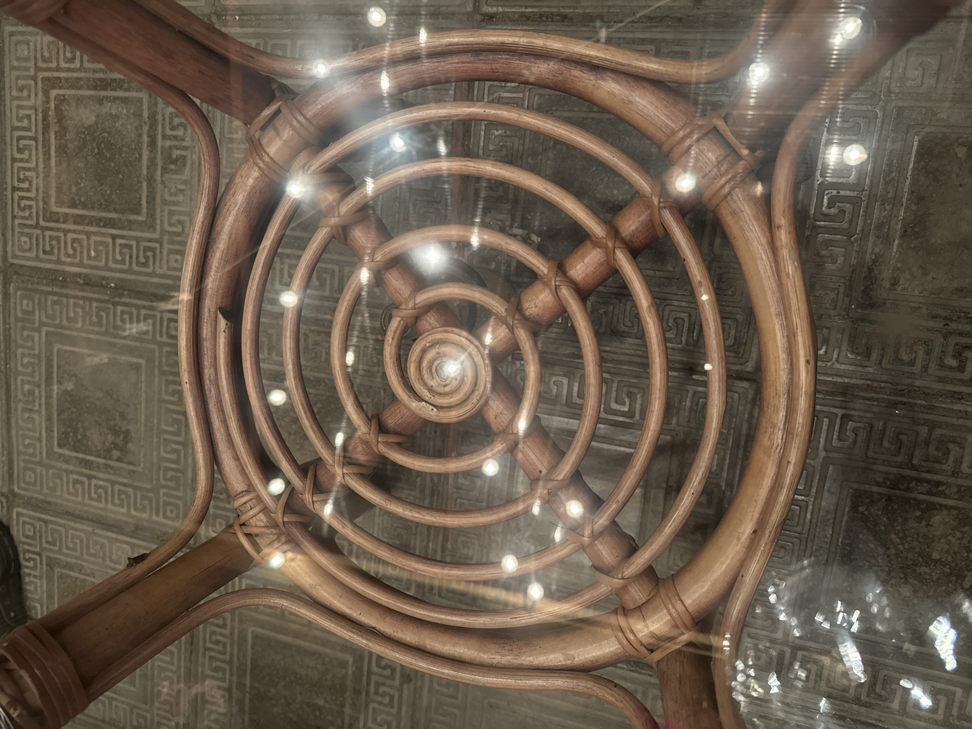 The image shows the underside of a rattan chair. The chair has a circular design with concentric rings of rattan, tied together with smaller pieces of rattan. The chair is placed on a tiled floor with a geometric pattern. The scene is likely indoors. The image does not provide any information about the location in Chuguyevka, Russia.