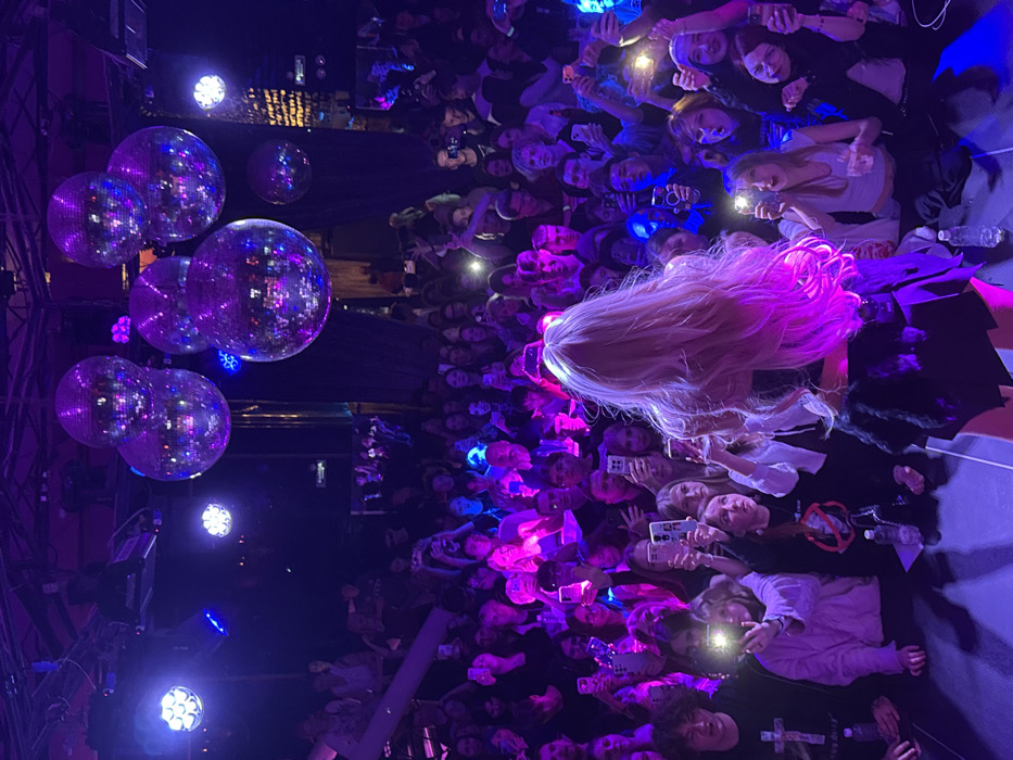 The image depicts an indoor live performance or concert event occurring at the Embargo Club (Embargo Villa) in Rostov-on-Don, Russia.
The primary subject is a performer, observed from behind, standing centrally on a stage. This individual has long, light-blonde hair and is wearing dark attire, appearing to consist of a top and a skirt or shorts. The performer is illuminated by bright stage lighting, casting a purple glow on their hair and outline. A plastic water bottle is visible on the stage near the performer's feet.
A dense crowd of several hundred individuals, primarily young adults and teenagers, occupies the space in front of the stage and extends into the background, including a visible upper-level balcony or seating area. Numerous audience members are holding up smartphones, actively recording or photographing the performance, with several phone screens and camera flashes illuminated, creating bright spots in the dark environment. Their gazes are directed towards the stage.
The venue's interior is dimly lit overall, with prominent stage lighting fixtures casting strong blue and purple hues across the stage and audience. Several large, spherical disco balls are suspended from the ceiling, reflecting light. The background reveals architectural elements of the club, including dark walls and structural beams. Another plastic water bottle is visible on the floor in the immediate foreground among the audience.