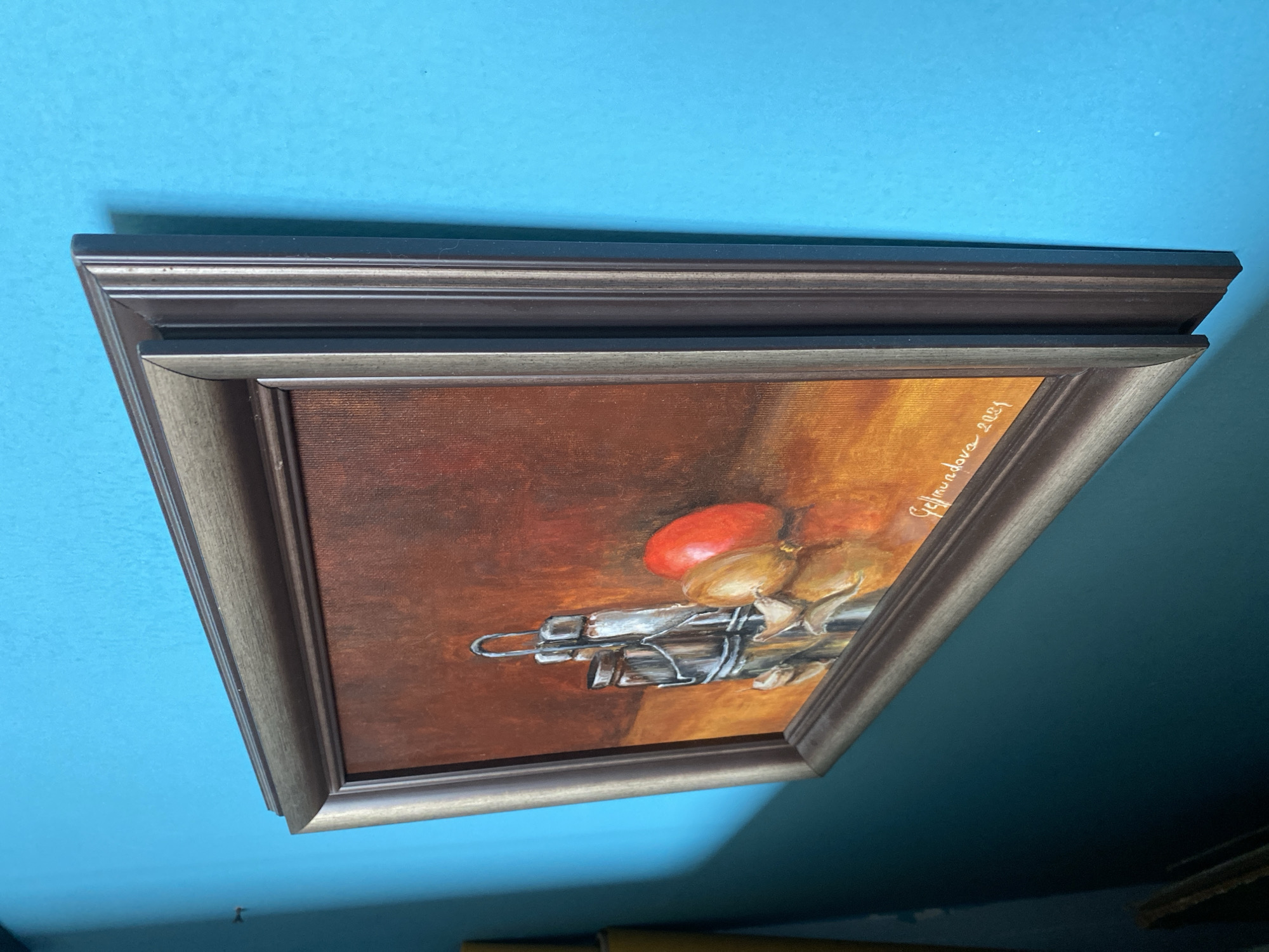A framed still life painting hangs on a bright blue wall. The painting depicts a salt and pepper shaker, a red tomato, and an onion. A signature and the year "2021" are visible in the bottom right corner of the painting. The wall is smooth and painted a vibrant turquoise. The lighting casts a subtle shadow to the left of the painting. The context of Bílina, Czechia, is not discernible from the image.