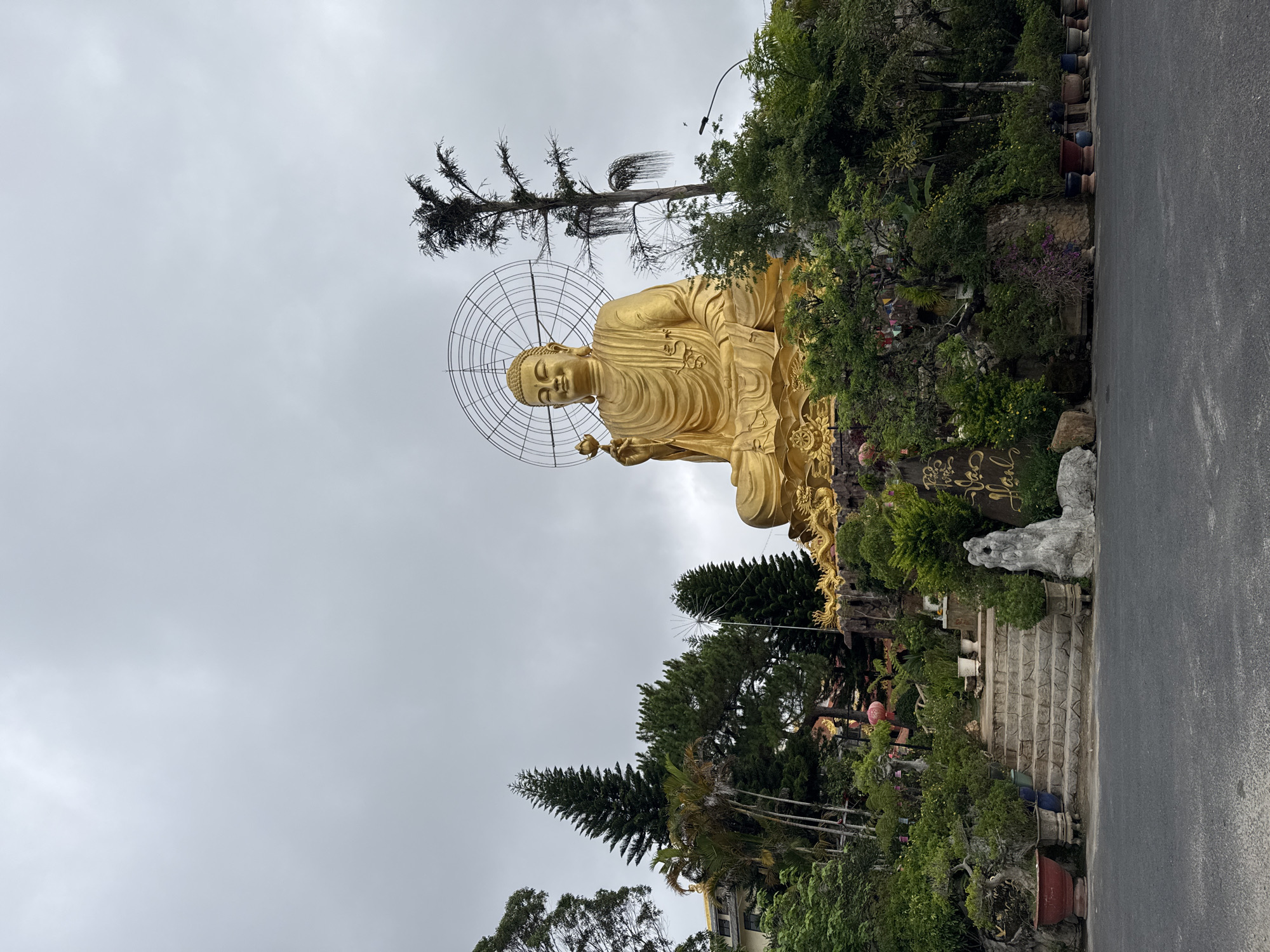 A sideways, low-angle shot captures a large, golden Buddha statue against an overcast sky in Lâm Viên, Đà Lạt, Vietnam. The statue, encircled by a metal aureole resembling a web, sits in a lotus position, holding an object in its right hand. It is surrounded by lush green trees and vegetation. A set of stone steps leads up to the statue. To the right of the steps is a gray, stone sculpture. A paved road runs along the right side of the image. The sky is a uniform gray, suggesting an overcast day. The scene is peaceful and serene.