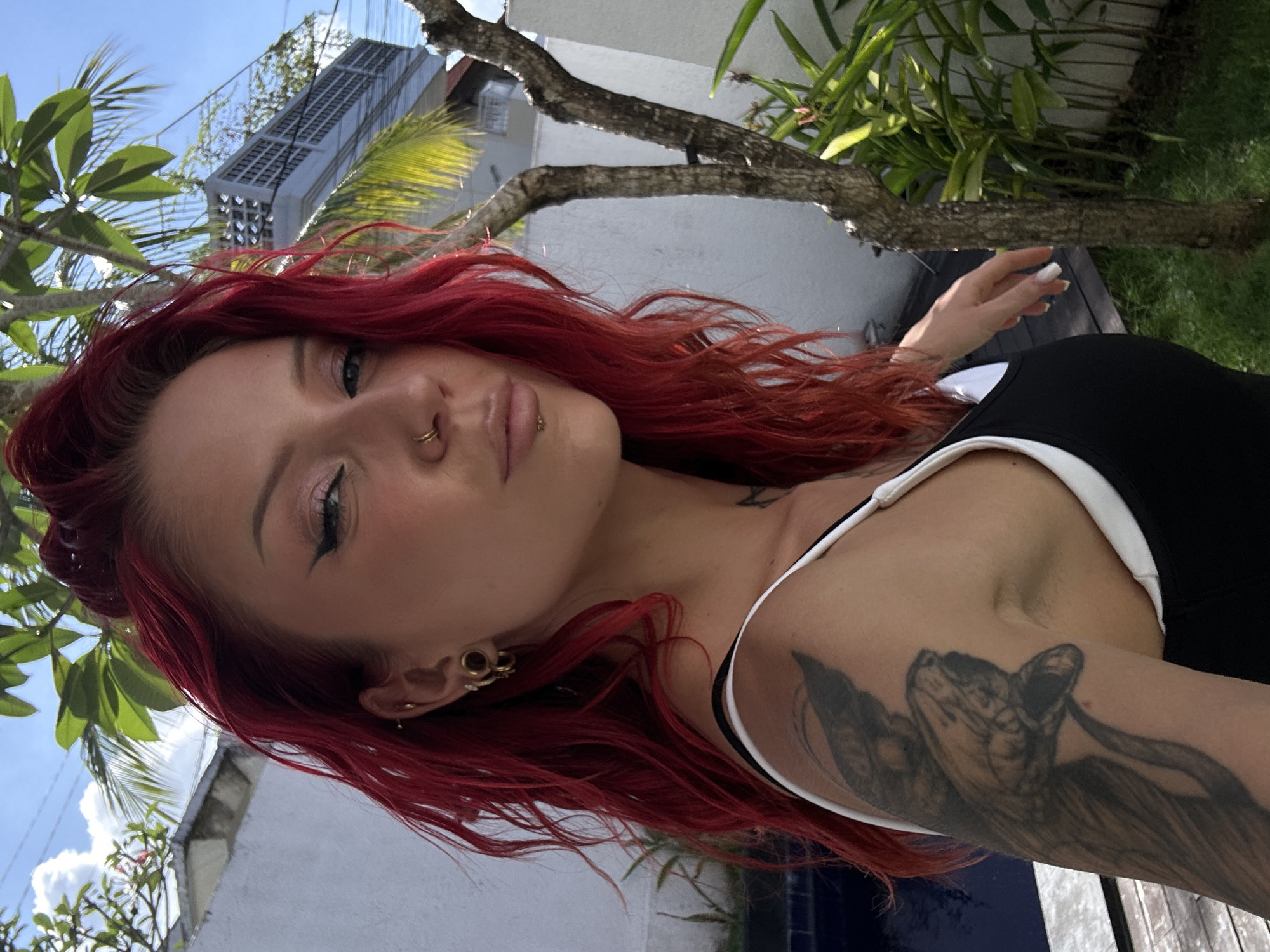 A young woman with bright red hair and multiple ear piercings is posing for a selfie outdoors in what appears to be a residential backyard in North Kuta, Indonesia. She is wearing a black and white tank top and has a tattoo of a snake on her left arm. The background shows a swimming pool, lush green foliage, a tree, and a building with a patterned facade. The sky is blue with some clouds, and the sun is shining.