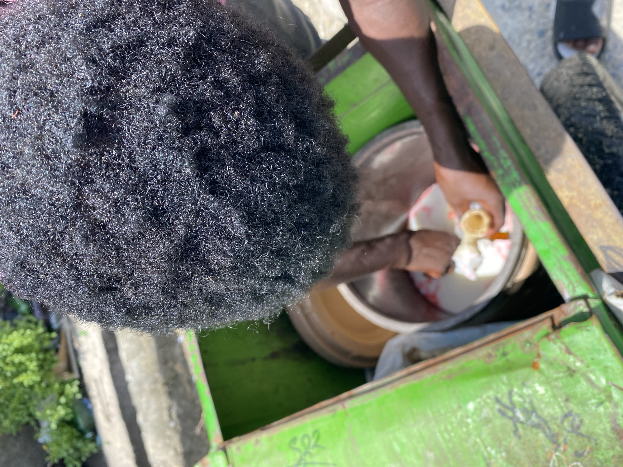 The image is a top-down shot that appears to be taken in the city of Unknown, Unknown. The primary subject is a person with a large, black afro hairstyle, who is standing next to what seems to be a green cart. The person's right hand is holding a cone filled with what looks like ice cream, inside a metallic container filled with white substances, presumed to be ice cream.
The cart, green in color, appears to be portable and likely used for selling the ice cream. A foot wearing a black slide is visible on the upper-right of the frame, along with a black tire, suggesting this is a mobile vendor. The ground appears to be concrete, and some greenery is visible in the lower-left corner, potentially indicating an outdoor location.