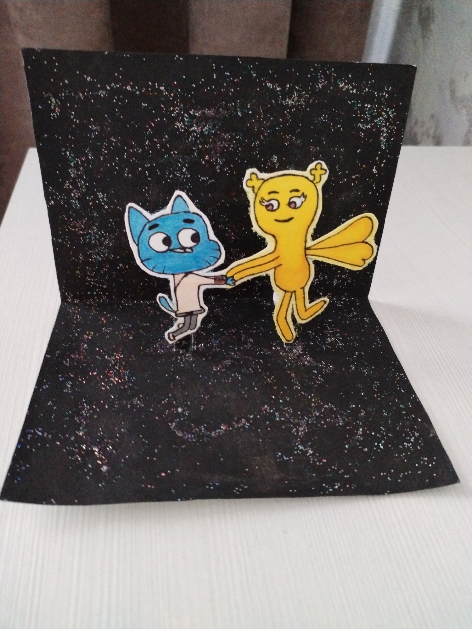 A handmade pop-up card featuring two cartoon characters, Gumball Watterson and Penny Fitzgerald from the animated series "The Amazing World of Gumball," is presented against a black background sprinkled with glitter to simulate a starry sky. The card is open, displaying a 3D effect with the characters positioned on the folded sections. Gumball, a blue cat wearing a white shirt, and Penny, a yellow creature with wings, are shown holding hands and appearing to dance or move together. The card is placed on a white, ribbed surface, likely a table, with a blurred background that suggests an indoor setting, possibly a home. The image captures the card from a slightly elevated, front-facing perspective. This scene is not located in Хідрынскі сельскі Савет, Belarus; it is an indoor, man-made object.