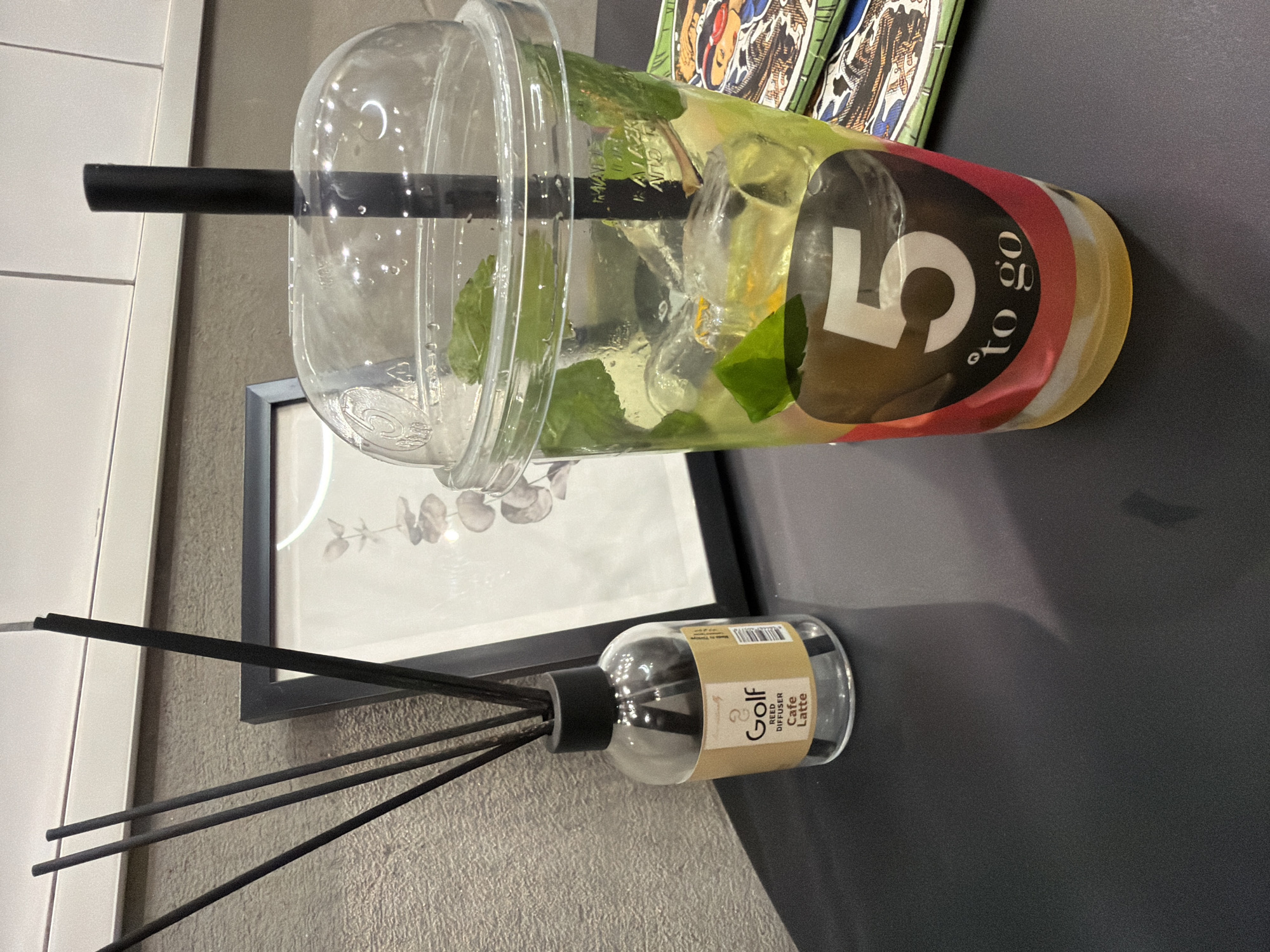 The image shows a close-up view of a black table with several objects. In the foreground, a clear plastic cup filled with a light-colored beverage, ice cubes, and mint leaves is prominently displayed. The cup has a clear plastic lid and a black straw. A sticker on the cup features a large white number "5" and the text "to go" in smaller letters. To the left of the cup, a small glass bottle with a black cap contains dark reeds, indicating it is a reed diffuser. A label on the bottle reads "Golf REED DIFFUSER Cafe Latte". Behind the diffuser, a framed picture with a light background and a botanical illustration is visible. The background wall has a textured gray surface and a white horizontal border. A stack of colorful items with artistic patterns is partially visible to the right of the cup. The lighting suggests an indoor setting. The location is not specified.