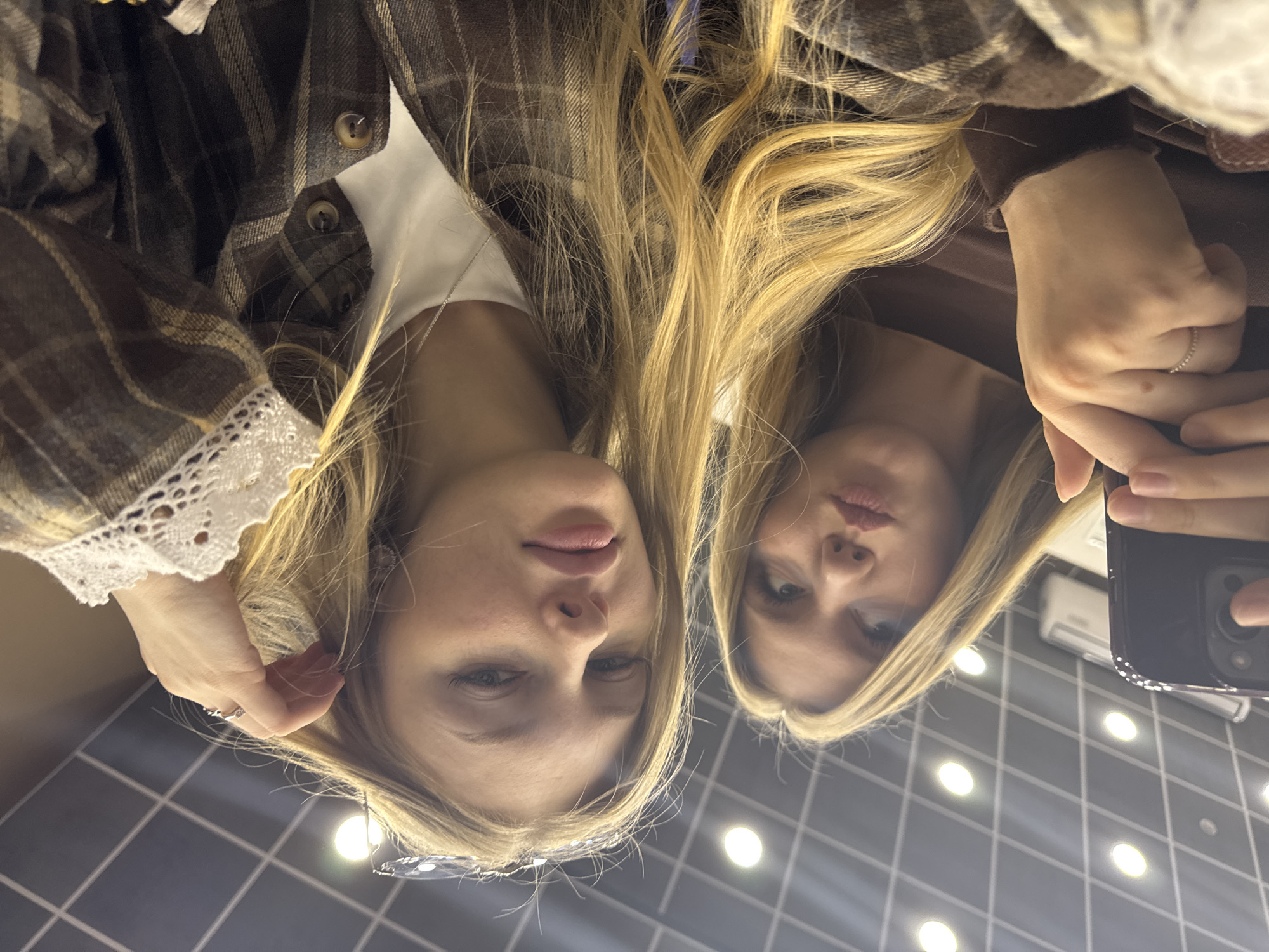 The image displays a rotated reflection of two young women, likely a mirror selfie. Their faces are prominently featured, appearing to look slightly downward from the top of the frame due to the image's 90-degree clockwise rotation. Both women have long, blonde hair and are wearing plaid shirts over lighter-colored tops.
The woman on the left wears a brown, tan, and white plaid shirt, revealing a white garment underneath with a distinct white lace-trimmed cuff on her sleeve. Her hand, adorned with a ring, is visible near her face. The woman on the right also wears a plaid shirt, layered over what appears to be a brown top. Her right hand, also featuring a ring, holds a dark-colored smartphone, which is partially visible in the reflection. Both women have subtle, neutral expressions as they look towards the reflective surface.
The background of the reflection reveals an indoor setting: a ceiling composed of dark gray or black square tiles with a bright white grid pattern. Several circular, bright white lights are embedded within these ceiling tiles, casting a clear illumination. A light-colored rectangular fixture, possibly an air vent or part of an air conditioning unit, is also visible in the upper right portion of the reflected background.
The activity taking place is the capturing of a self-portrait or group photo using a reflective surface, likely a mirror, in an indoor environment in Perm, Russia. The artificial lighting suggests no specific time of day. No visible text is present in the image.