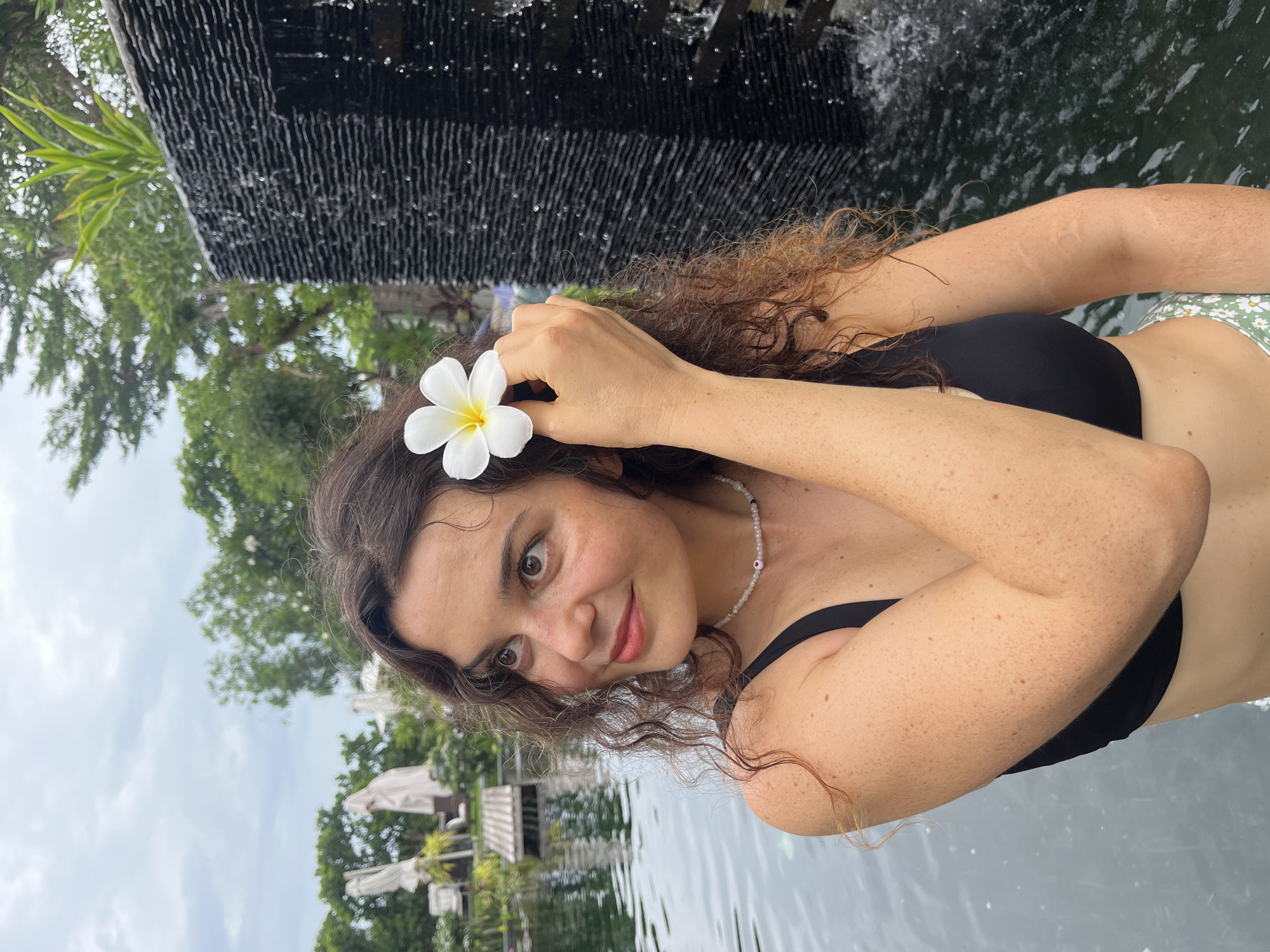 A young woman with curly brown hair and freckles is holding a white plumeria flower behind her ear while sitting by a pool with a waterfall feature. She is wearing a black bikini top and a necklace. The background shows greenery and umbrellas, suggesting a resort or hotel setting in Mũi Né, Vietnam. The lighting is overcast, indicating a cloudy day. The water in the pool is dark and reflective, and the waterfall creates a textured effect with cascading water.