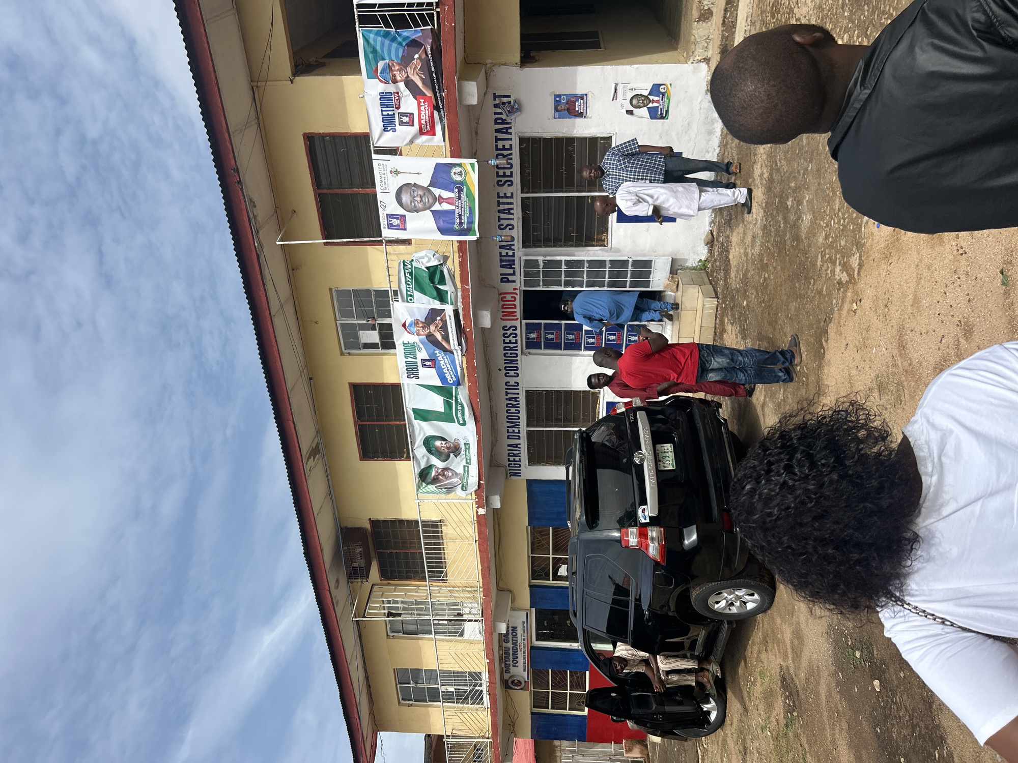 Here is a concise description of the image:
**Setting:** A two-story building is identified as the Nigeria Democratic Congress (NDC), Plateau State Secretariat.
**Location:** This building is likely situated in Jos, Nigeria.
**Subject:** Political banners featuring candidates' faces and names such as "SABON ZANCE," "OBADIAH DAMWAGHA," "CEOFFREY BOTTSON," and "SOMETHING NEW" decorate the building's facade. The Dayyabu Gawu Foundation is also featured on the building.
**Objects:** A black Toyota Land Cruiser Prado is parked in front.
**People:** A small group of people is visible, some interacting near the car. One is wearing a red shirt and jeans. A man in a white traditional outfit stands near the entrance. Two others are dressed in black, their heads partially visible. Another person in a white shirt is situated in the foreground.