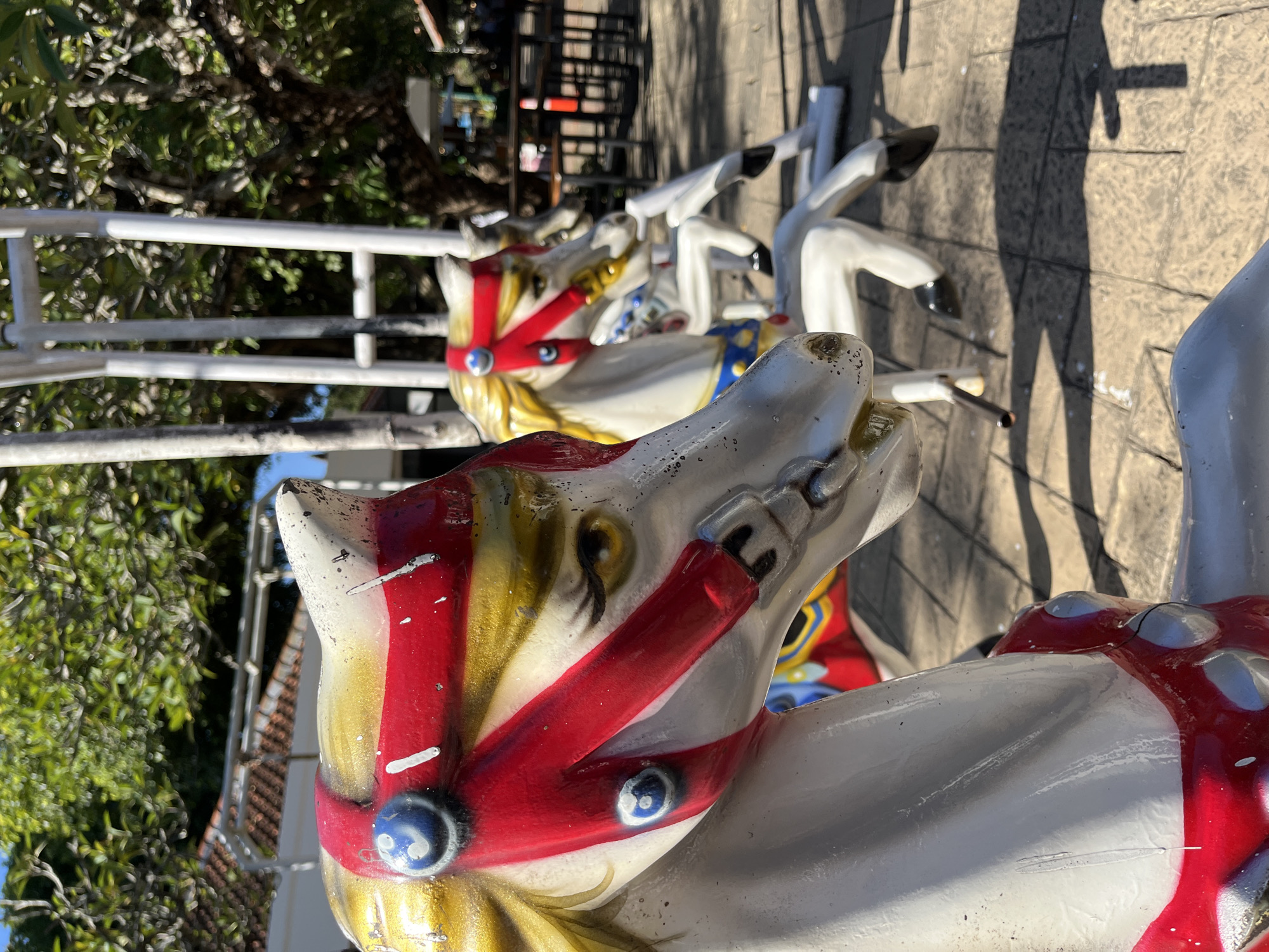 The image shows a carousel horse, painted white with a red harness and gold accents, with another horse partially visible in the background. The horses are outside on a stone surface near trees. The location is Unknown, Indonesia.
