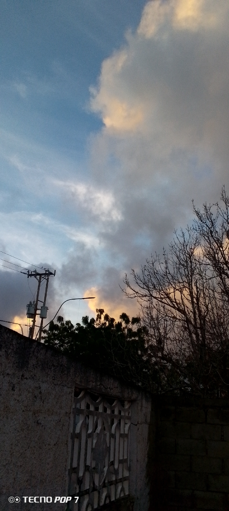 The image captures a twilight scene in Altagracia, Venezuela, characterized by a dramatic sky and the silhouette of urban elements. The sky is a blend of blues, grays, and oranges, with clouds illuminated by the setting sun. Prominent in the lower left is a utility pole with transformers and overhead wires, typical of urban infrastructure. Adjacent to it, a wall with a decorative metal grate over a window or opening is visible. To the right, a bare-branched tree extends upwards, its dark silhouette contrasting with the colorful sky. The overall impression is one of a calm, yet evocative, end-of-day vista in a residential or semi-urban area. The bottom left corner displays a watermark "TECNO POP 7".