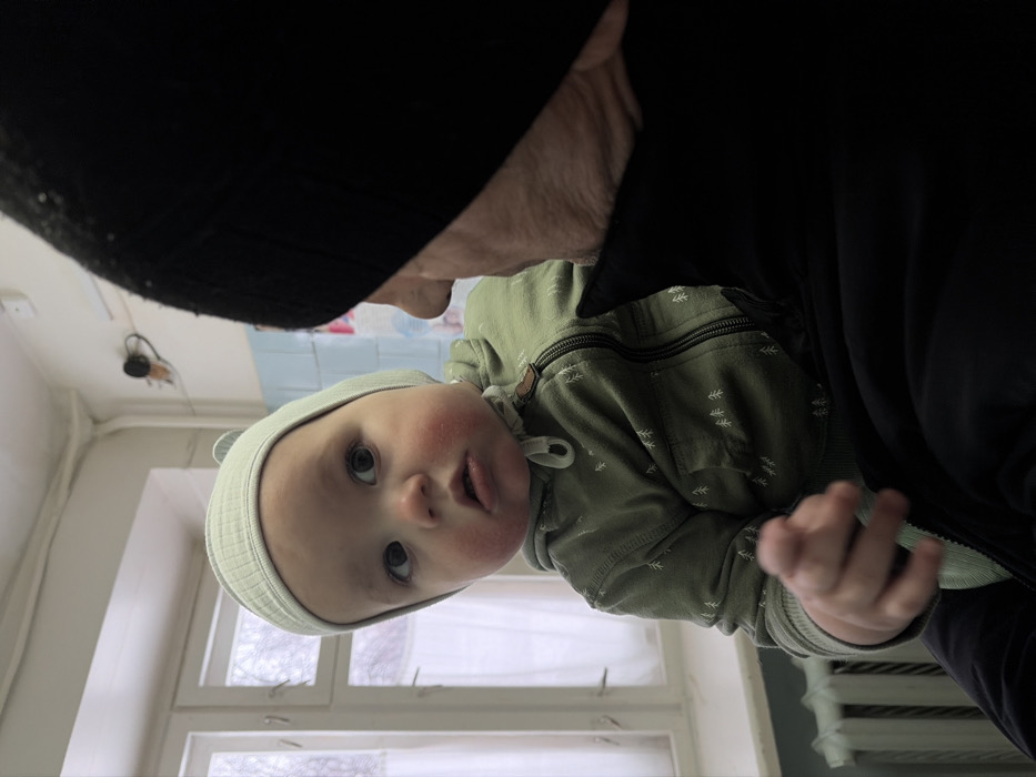 A baby with rosy cheeks and a knit hat looks up towards an adult wearing a black hat and jacket. The baby is wearing a green hooded sweatshirt with a pattern of white trees. The background shows a window with white frames and curtains. The photo is taken from a tilted perspective, with the ceiling and the top of the adult's head at the top of the frame.