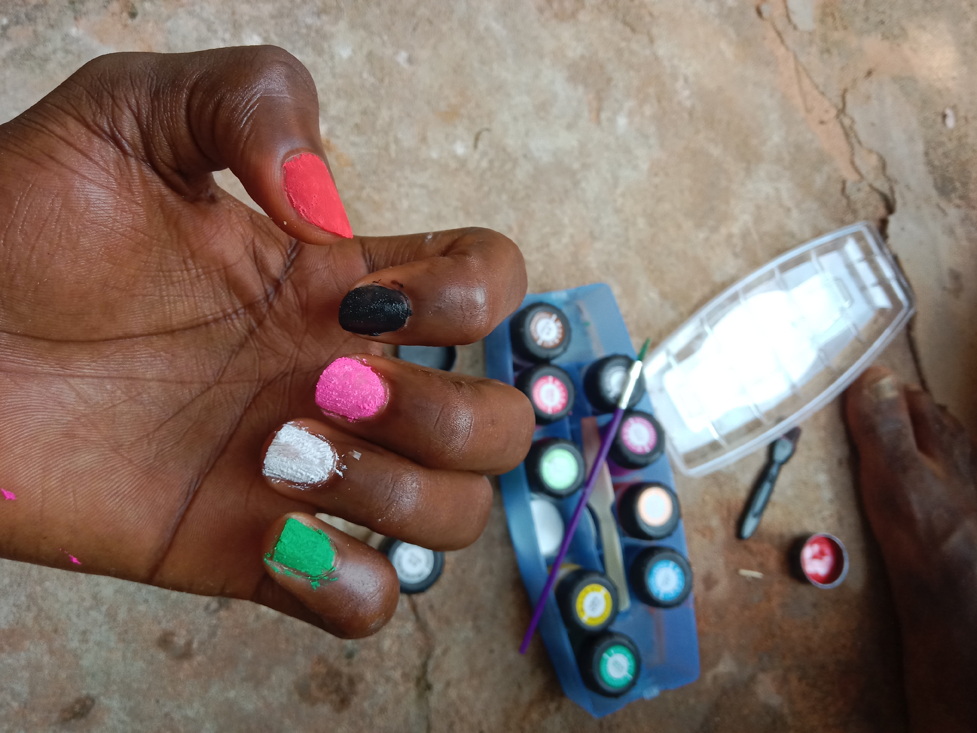 The image depicts a person's hand with painted nails. The nails are painted in different colors: red, black, pink, white, and green. Next to the hand, there's a palette of paints with various colors and painting tools. The background appears to be a textured surface, possibly concrete or stone. The location is unknown.
