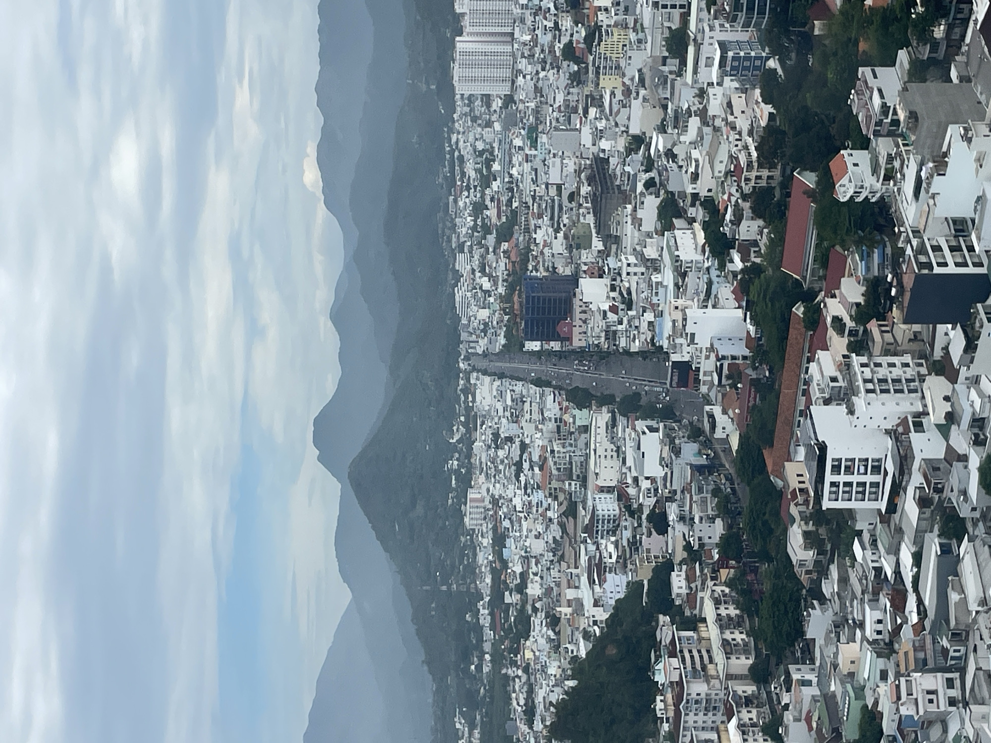 The image captures a panoramic view of Nha Trang, Vietnam, from an elevated vantage point. The city is densely populated with a mix of low-rise buildings and several high-rise structures, including apartment complexes and hotels. A wide road, likely a main thoroughfare, bisects the urban landscape, with traffic visible. Greenery is interspersed throughout the city, with trees and small parks dotting the skyline. In the background, a range of lush, green mountains forms a dramatic backdrop against a hazy, overcast sky. The overall impression is one of a vibrant coastal city nestled amidst natural beauty.