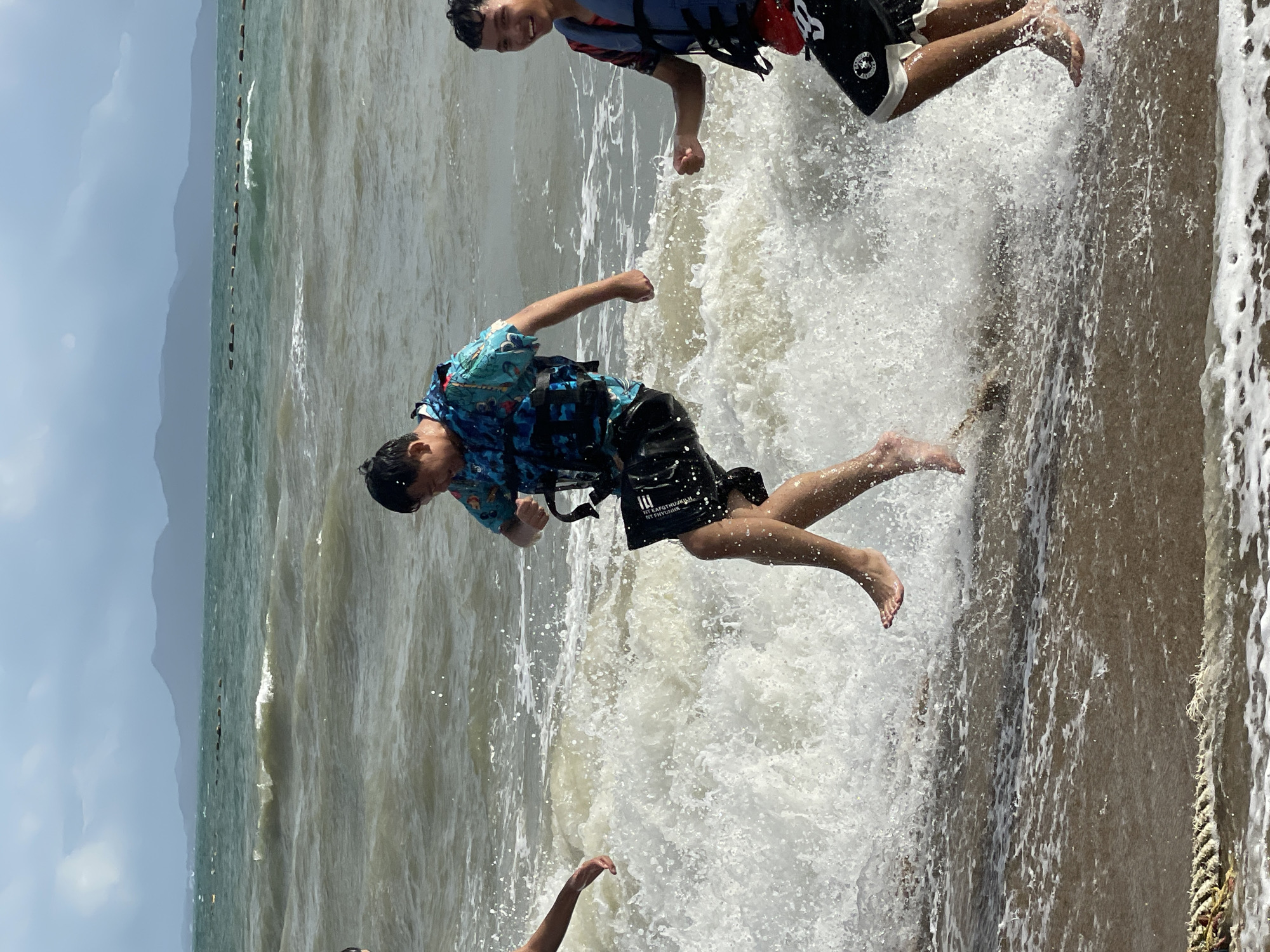 The image depicts three young males wading in the ocean at Pattaya Beach, Thailand. The water is choppy with breaking waves. The person in the center, wearing a blue and black life vest over a colorful shirt and black shorts, is mid-stride, with one leg raised as if jumping. To the right, another young male, wearing a red and blue life vest over a patterned shirt and black shorts with white trim, is smiling and looking towards the camera. His left hand is clenched. To the left, partially visible, is a third individual reaching out with their left hand. The sky is overcast with a range of mountains visible in the distance. A floating buoy line stretches across the water. The shore is covered in wet sand with some foam and debris.