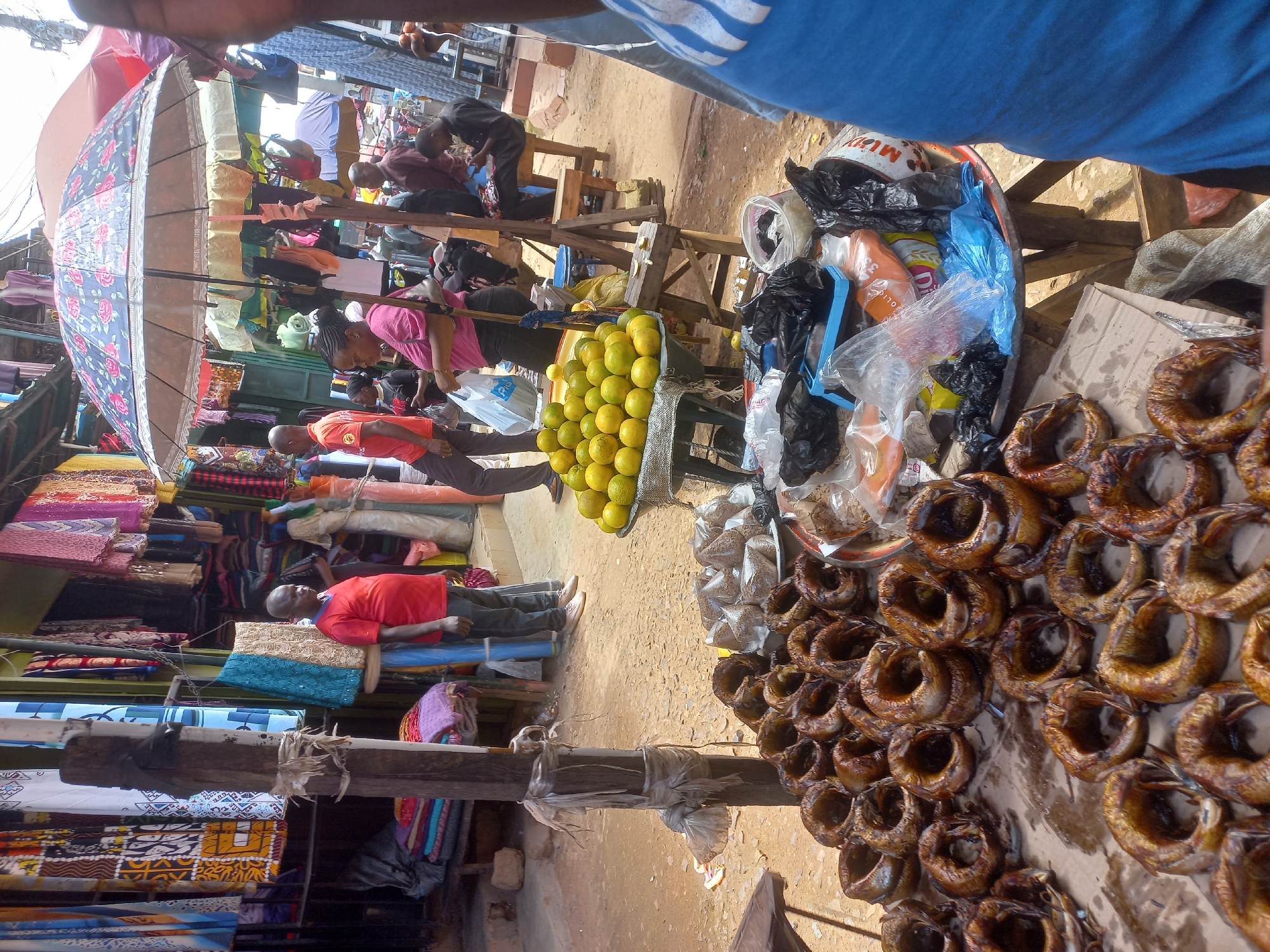 The image depicts a bustling outdoor market scene in Jos, Nigeria. In the foreground, a large display of smoked fish, arranged in a circular pattern on a brown surface, is prominently featured. Adjacent to the fish are bags of spices or grains, and further to the right, a metal bowl holds various items, including plastic bags, a blue tray, and what appears to be packaged food.
In the mid-ground, a table displays a large quantity of oranges. Behind this, various market stalls are visible, with colorful fabrics and clothing hanging from them. Several people are present, including a man in a red shirt standing near the fabric stalls, and others browsing or working. An umbrella with a floral pattern provides shade over a section of the market.
The ground is sandy and uneven, typical of an outdoor market. The overall impression is one of vibrant commerce and daily life in a Nigerian city.