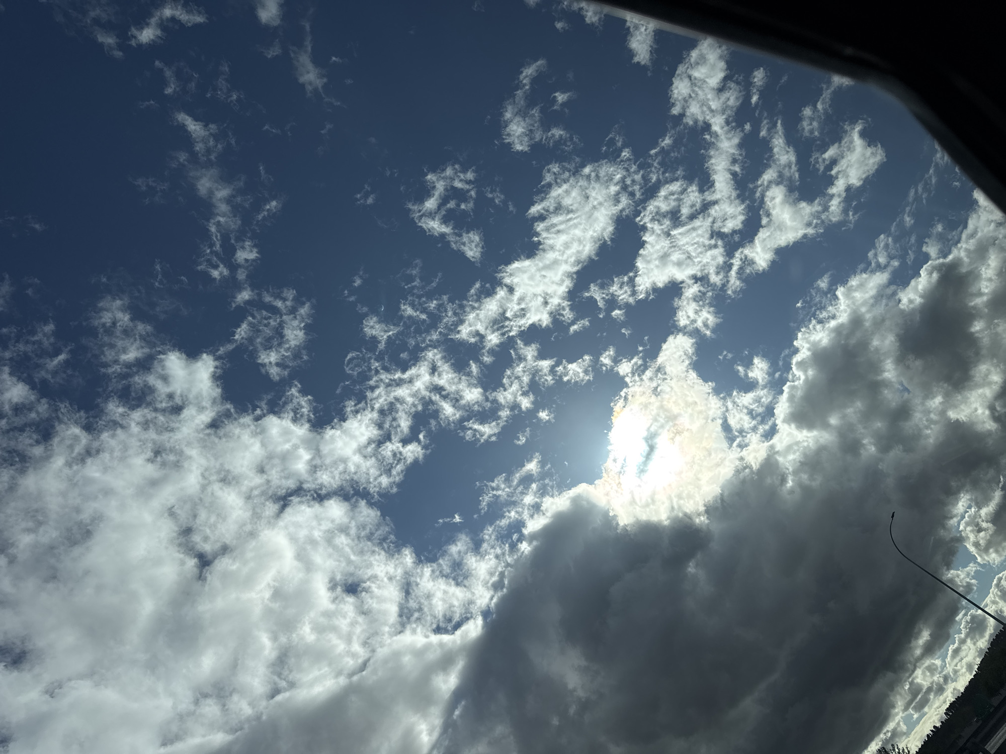 The image shows a view of the sky from an unknown location in Unknown, Unknown. The sky is blue with scattered white and dark gray clouds. The sun is visible behind some of the clouds, creating a bright glare. A lamppost and trees are visible in the bottom right corner. A small section of a car's roof is visible in the top left corner.