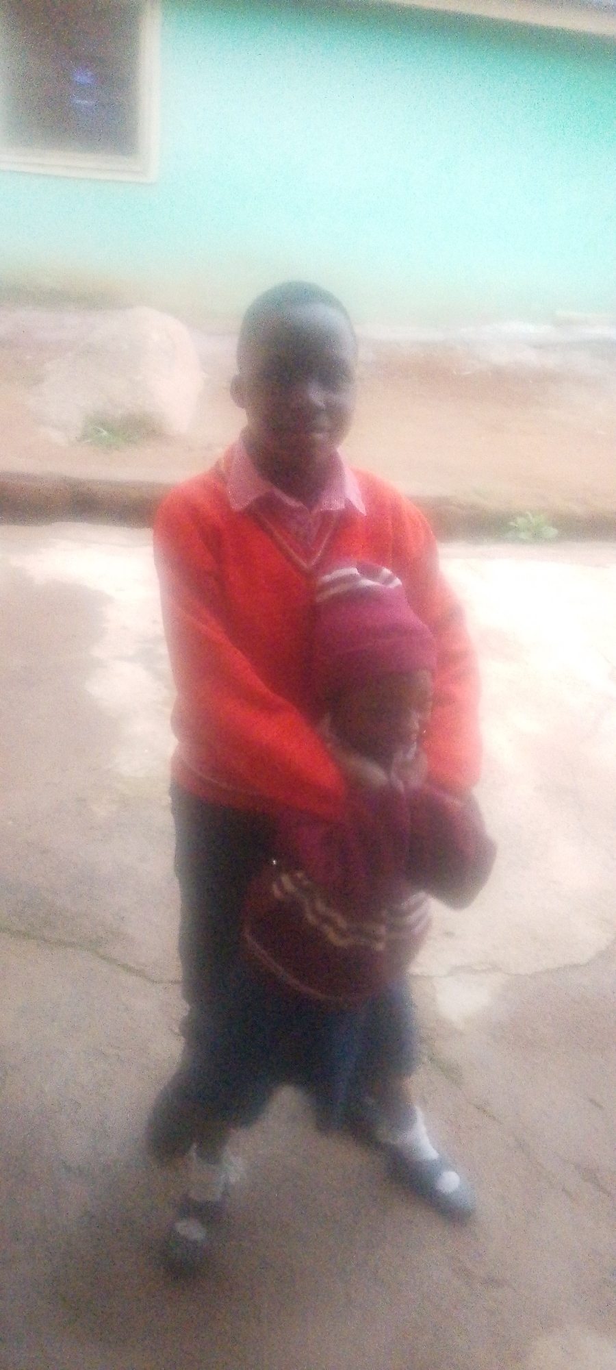 An overexposed, blurry, full-body shot shows two individuals, likely children, standing outdoors. The person in the foreground, on the left, is taller and wearing a red sweater over a collared shirt and dark pants. They are holding a smaller child in their arms. The smaller child, partially obscured, is wearing a striped sweater and a maroon hat. The background suggests an outdoor setting with a light blue wall, a window, and a concrete or dirt ground. The overall image quality is poor, with significant blur and noise.