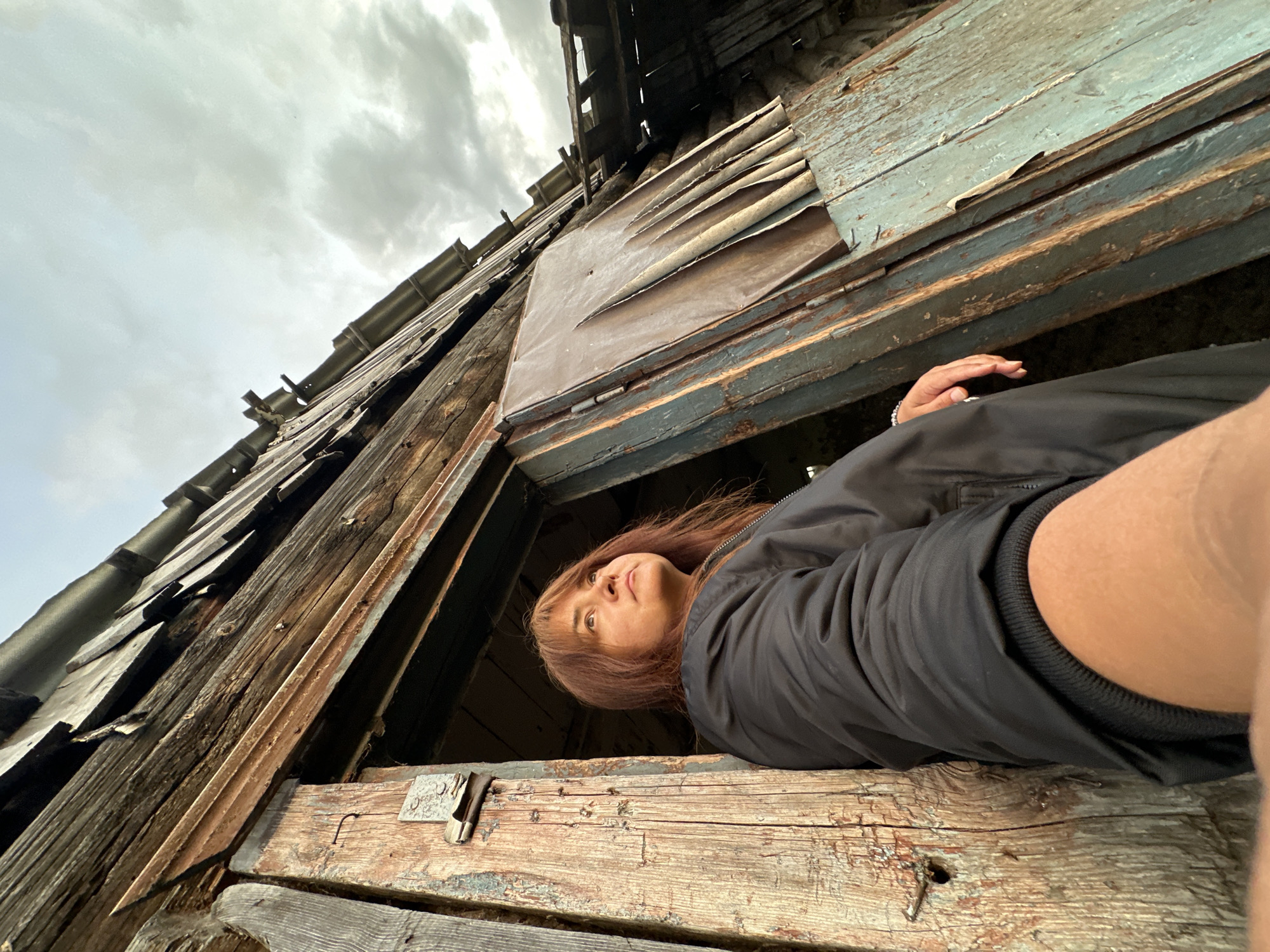 A woman with reddish-brown hair stands in a dilapidated wooden doorway, looking upwards and to the right. She is wearing a black jacket. The doorway is part of an old, weathered wooden structure with a partially collapsed roof made of uneven wooden planks. The wood of the structure is heavily weathered, with peeling paint and visible nail heads. The sky above is overcast with grey clouds. The perspective is a low-angle shot, emphasizing the dilapidated nature of the building and the upward gaze of the woman. The scene appears to be outdoors in a rural or abandoned setting.
