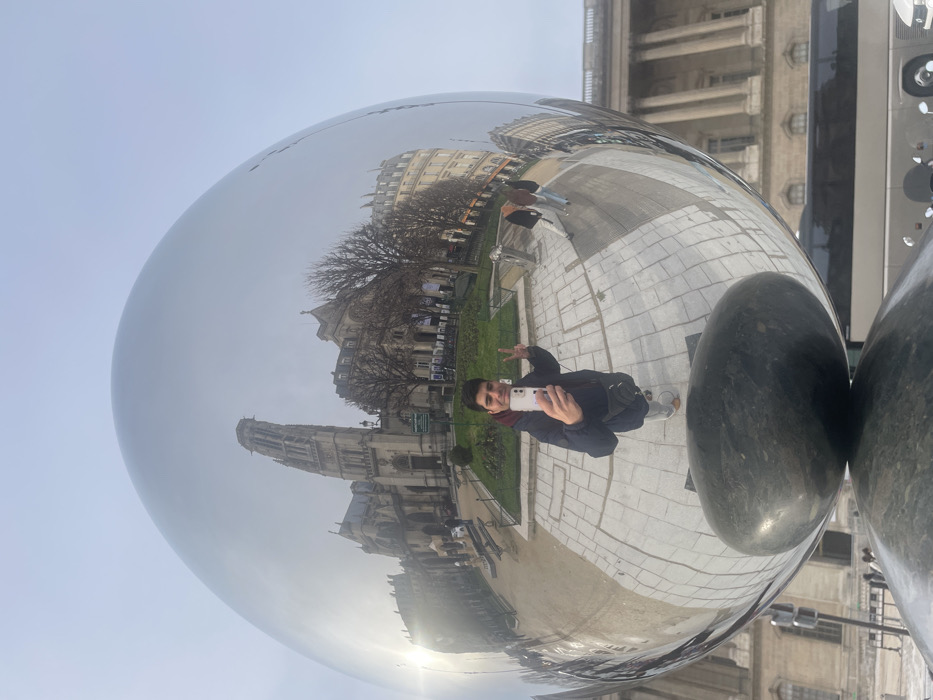 The image displays a reflective, egg-shaped stainless steel sculpture acting as a convex mirror, situated at the Place du Louvre in Paris, France. A dark, polished stone element forms the base of the sculpture. A male subject, centered in the reflection, is taking a selfie with a light-colored smartphone held in his right hand, while his left hand forms a peace sign. He is dressed in a dark puffer jacket, light trousers, and light shoes.
The reflection reveals the surroundings: prominent in the background is the Saint-Germain l'Auxerrois church, identifiable by its distinctively tall, ornate bell tower and gothic architecture. Adjacent to the church, typical Haussmannian-style buildings with multiple stories and classic Parisian facades are visible. A public green space with sparse trees, consistent with winter or early spring, and paved pathways separates the church from the immediate foreground. Two additional, less distinct figures are reflected walking on a paved area to the right of the main subject. The sky is overcast and bright. In the lower right portion of the reflection, a silver tour bus is partially visible, along with the classical columns of a large building, consistent with the Louvre Palace, across the street from the church.