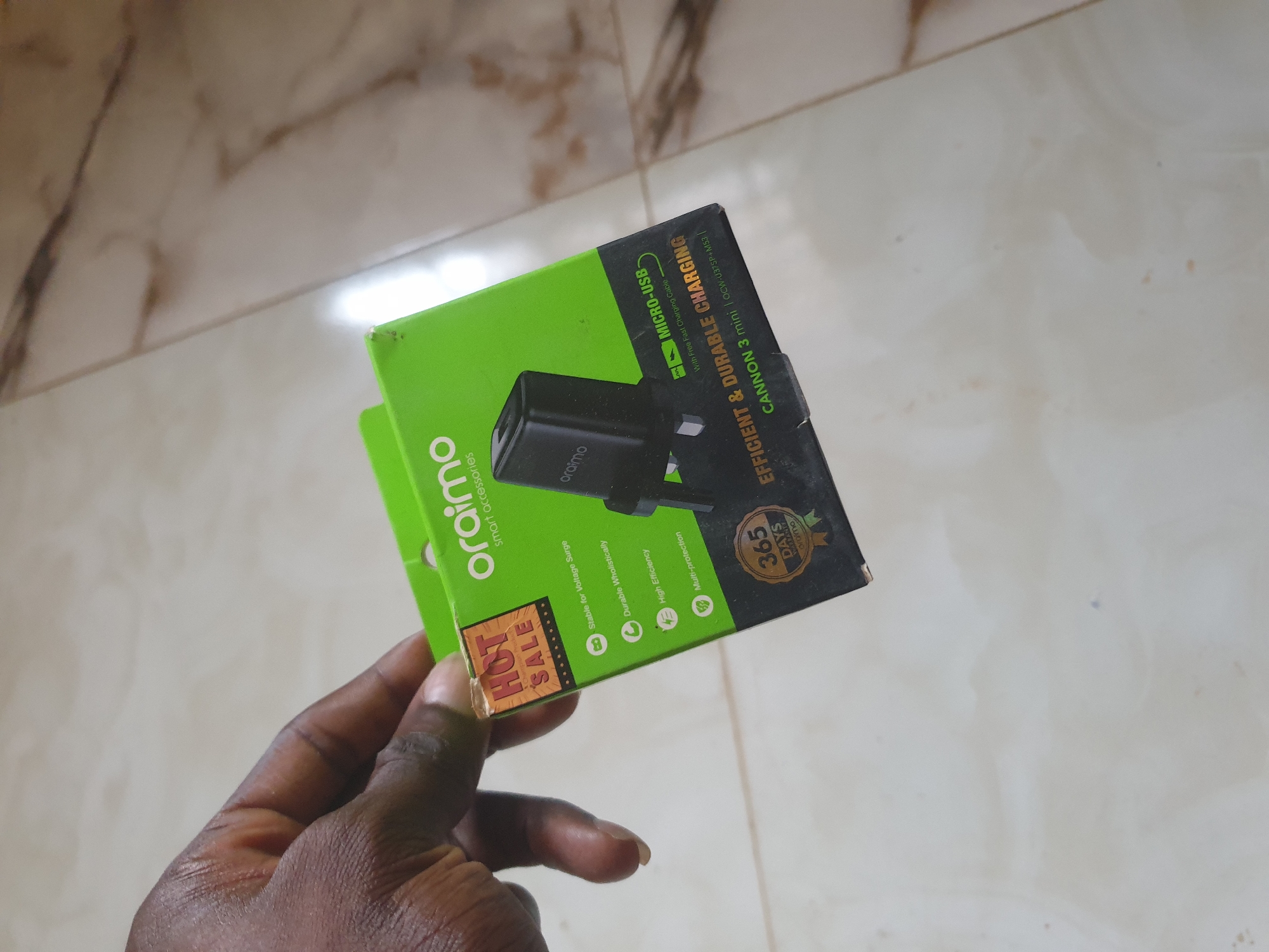 A person's hand is holding the corner of a bright green and black box for an Oraimo smart accessory. The box is oriented at an angle, with the front panel clearly visible. The "HOT SALE" sticker on the left side of the box is orange and red with white lettering. The Oraimo logo is printed in white on the upper part of the box. Below the logo, the text "smart accessories" is visible. A black electrical plug is depicted on the front of the box. Several features are listed with icons: "Stable for Voltage Surge," "Durable Wholistically," "High Efficiency," and "Multi-protection." At the bottom of the box, a circular emblem reads "365 DAYS WARRANTY" and "Oraimo." Further text at the bottom states "EFFICIENT & DURABLE CHARGING" and "CANNON 3 mini | OCW-U375P+M53." The background is a light-colored, tiled surface.